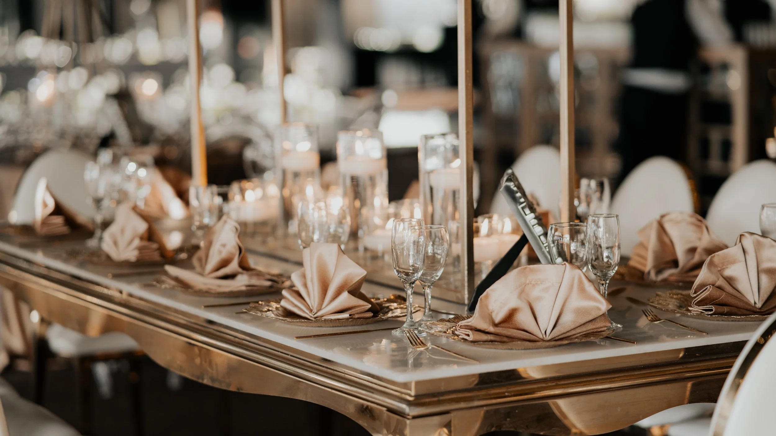 Mesa decorada de manera elegante con copas, cubiertos, platos, ánforas con velas, decoraciones doradas y servilletas dobladas en forma de flor para evento formal