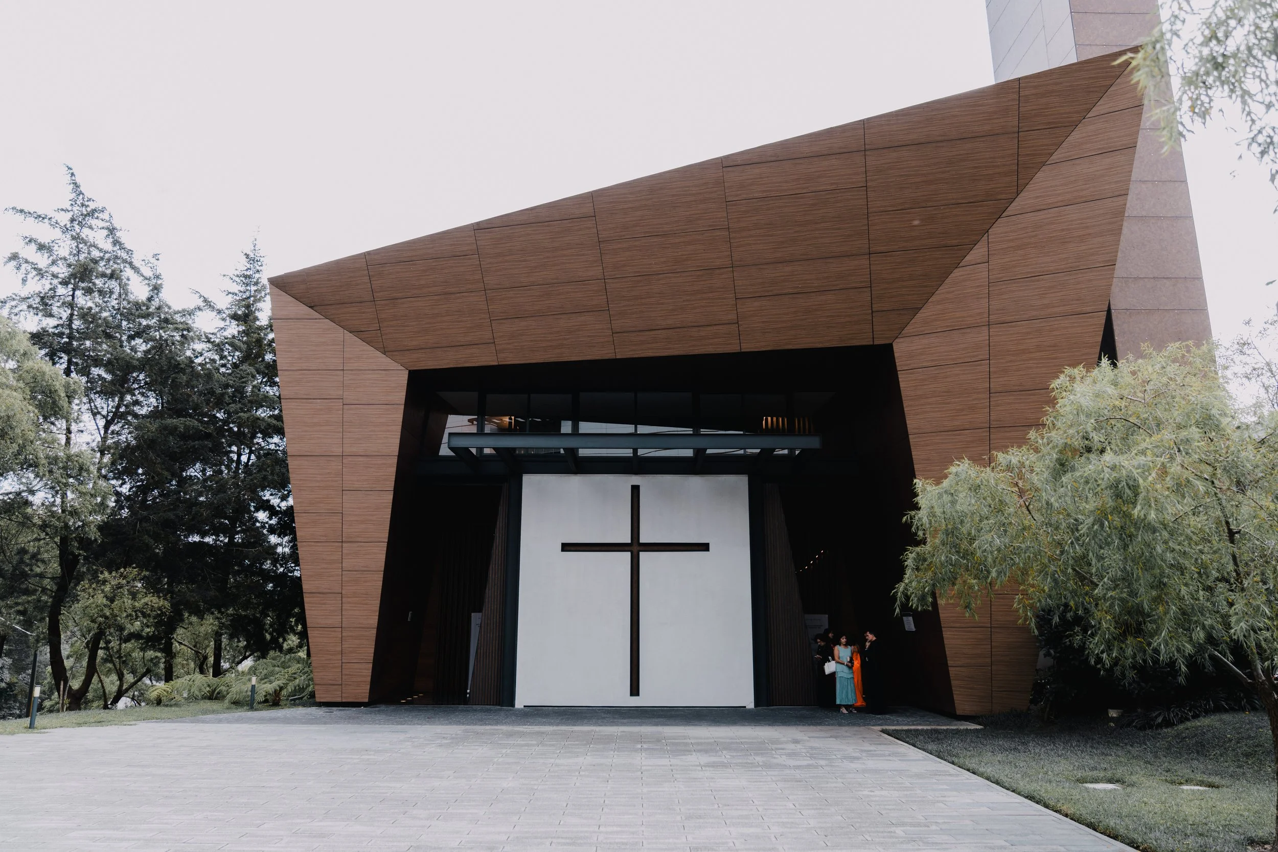 Fachada de una iglesia moderna con estructura de madera y una gran cruz en la entrada, rodeada de árboles.