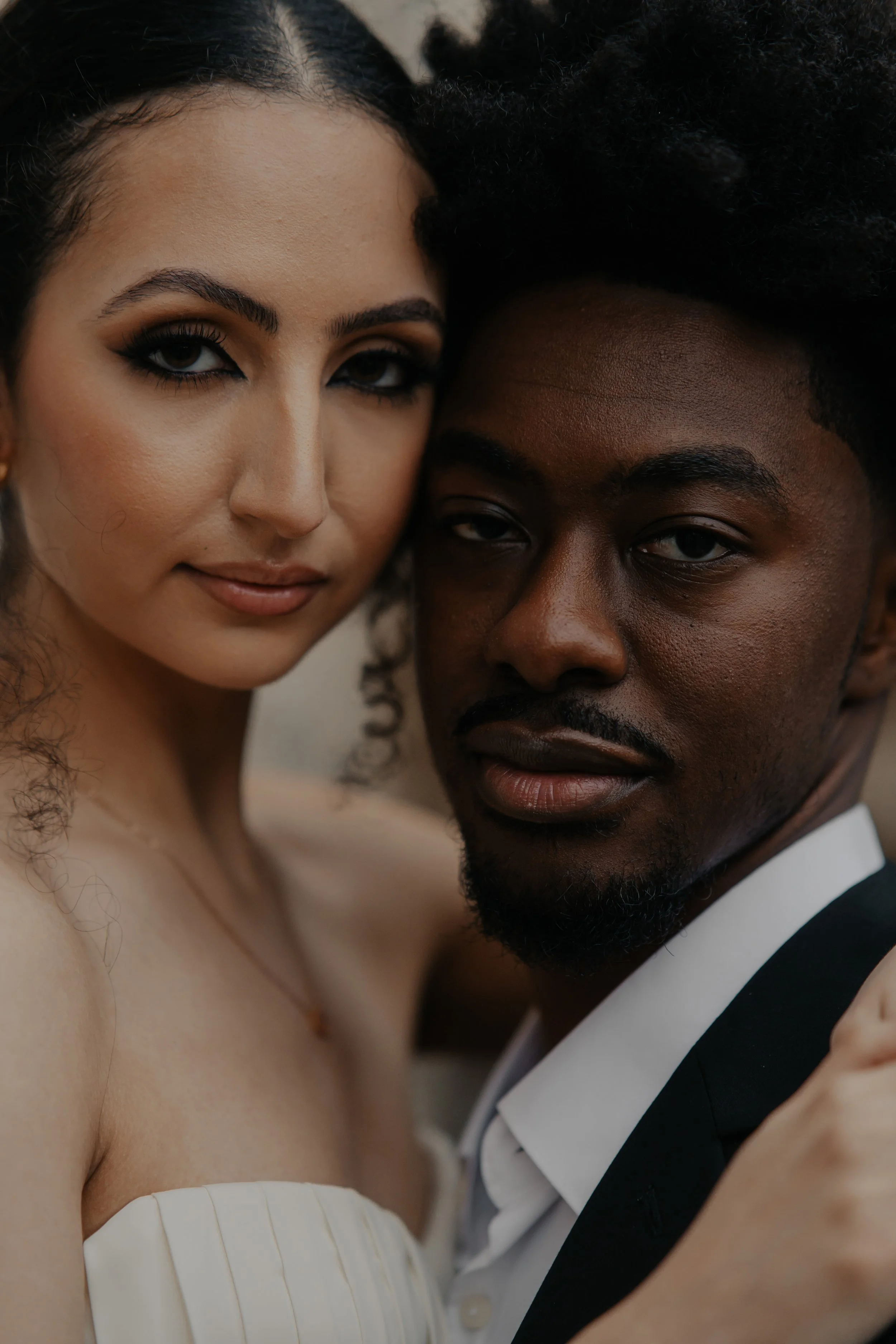 Una pareja joven en un retrato cercano, la mujer con maquillaje armonioso y cabello rizado, el hombre con barba y cabello rizado, ambos con expresión serena y mirada directa a la cámara.
