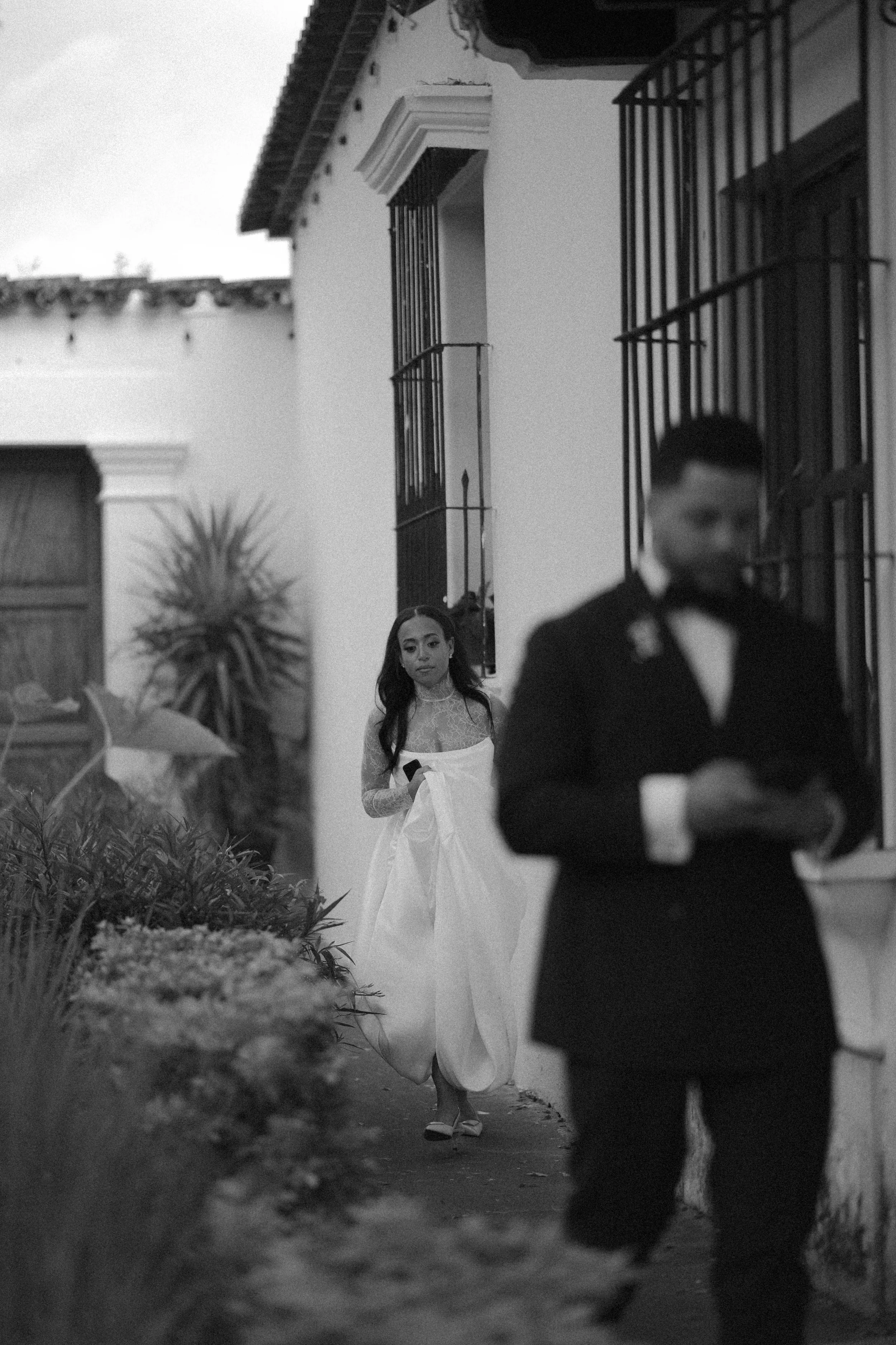 Una mujer vestida con un vestido blanco y un hombre con un traje negro en una escena en blanco y negro, en lo que parece ser una ceremonia o evento formal, en un entorno exterior con plantas y un edificio colonial.