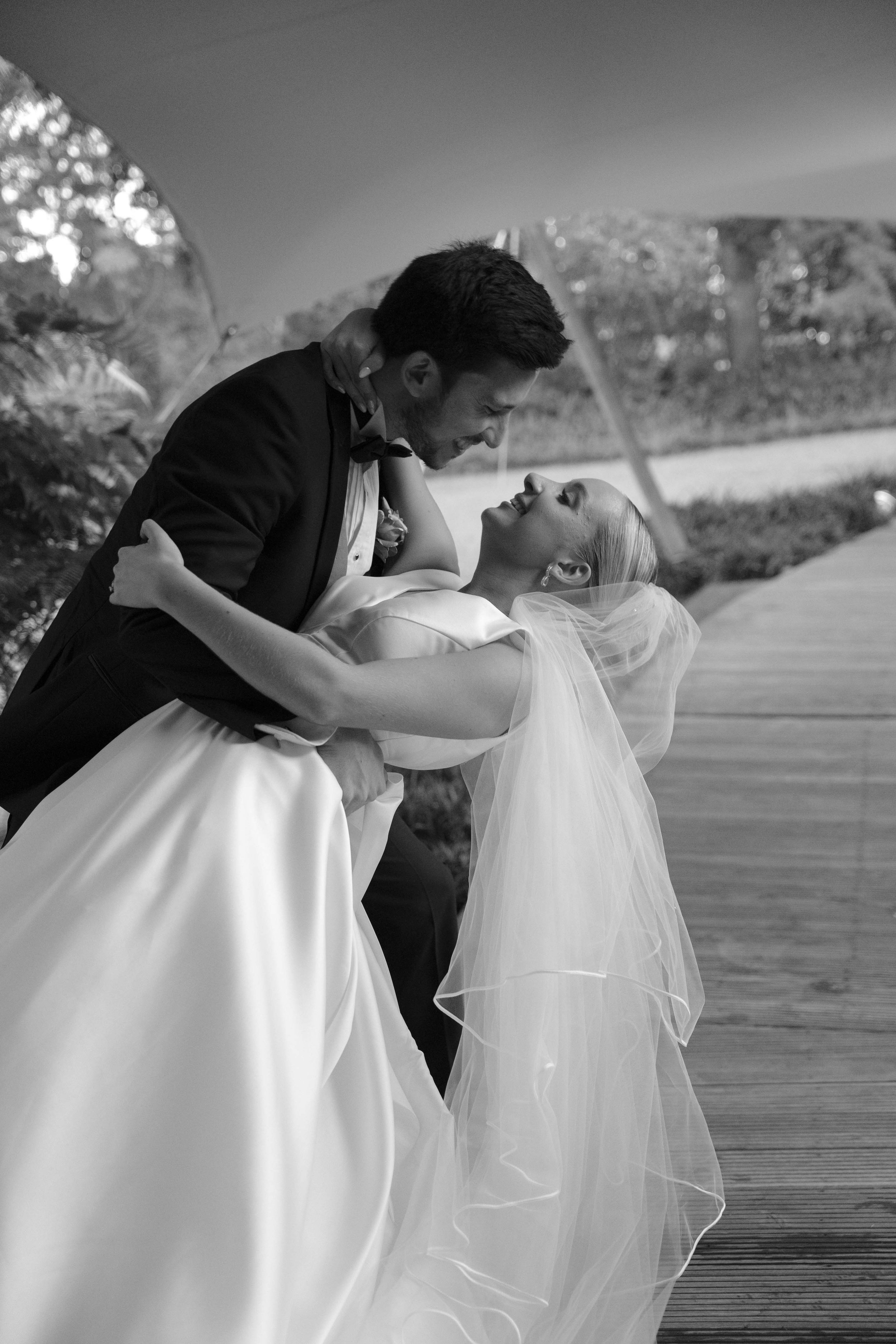 Pareja de recién casados bailando en exteriores, el hombre viste saco negro y la mujer lleva vestido de boda y velo, ambos sonrientes y felices.