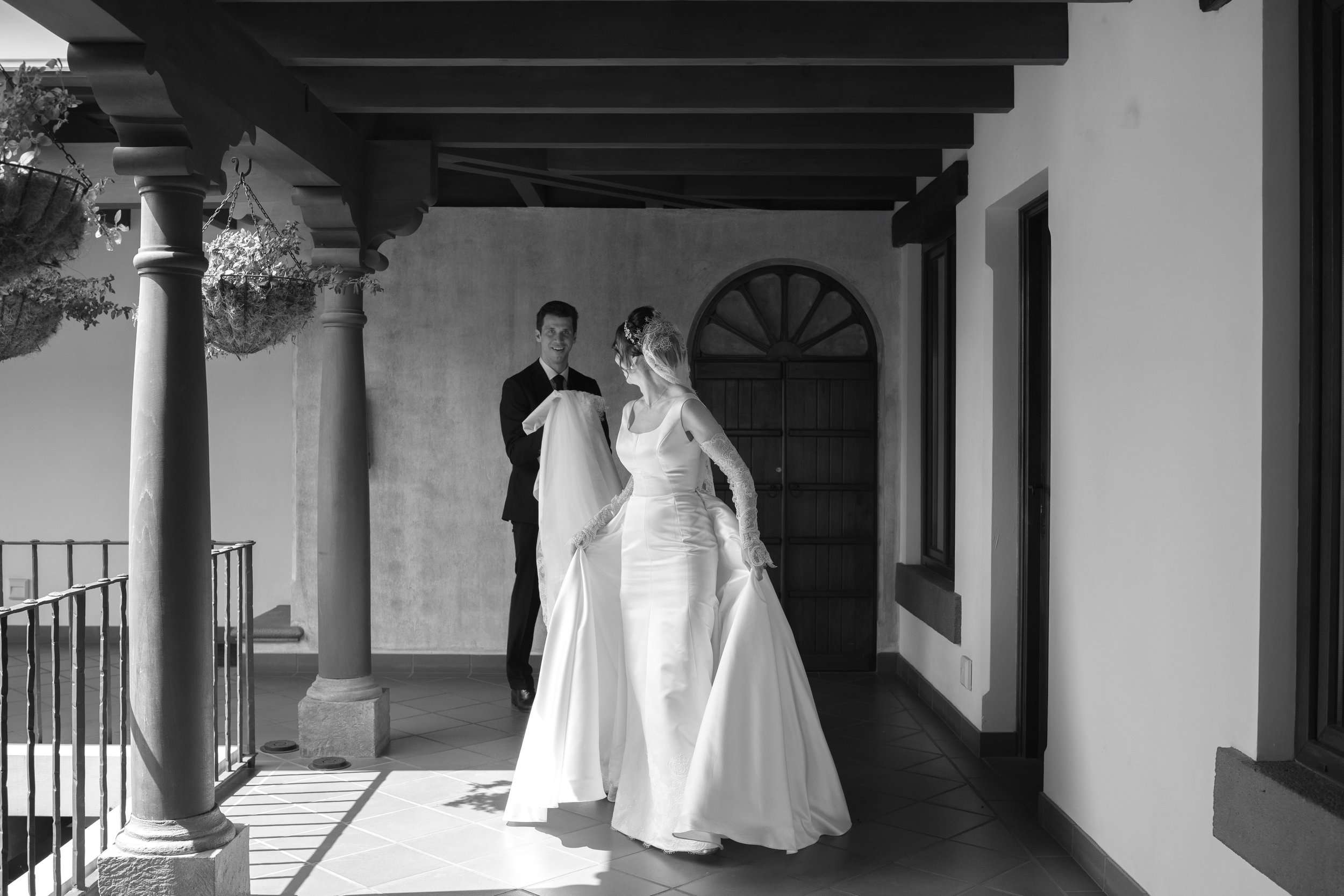 Pareja de novios en vestido de novia y esmoquin en balcón, con fondo de puertas y ventanas de madera.