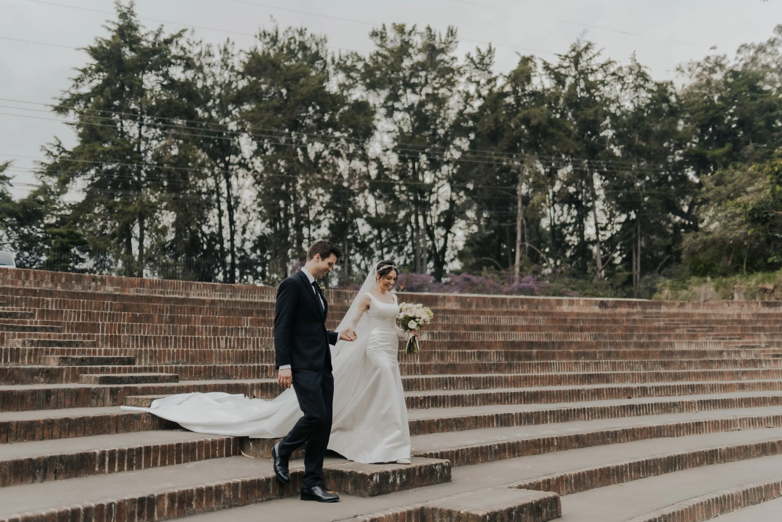 Una pareja de recién casados bajando por unas escaleras de ladrillo. La novia lleva vestido blanco y sostiene un ramo de flores, el novio viste traje oscuro.