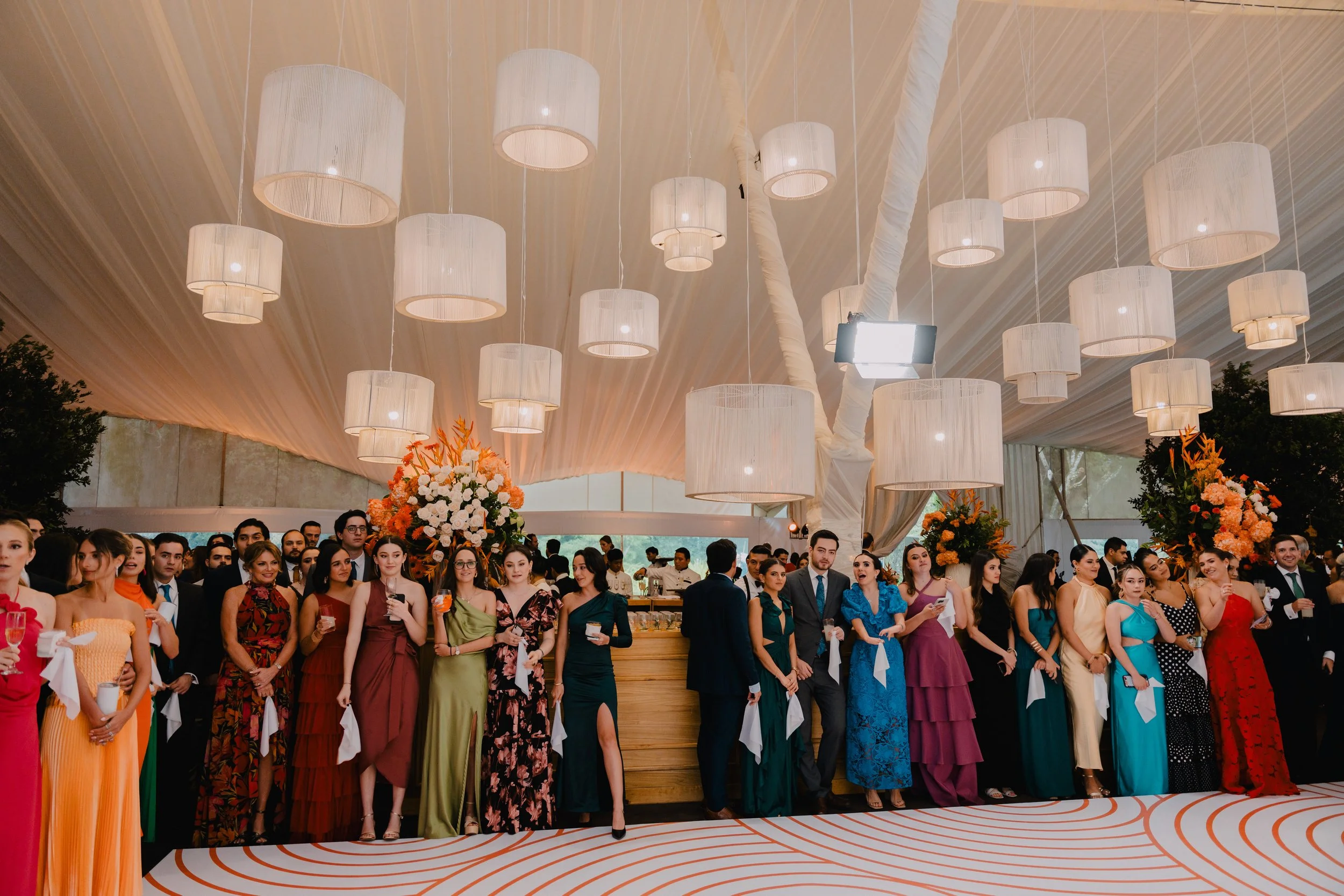 Grupo de personas en una recepción o fiesta, vestidas elegantemente, en un salón decorado con flores y lámparas grandes en el techo.