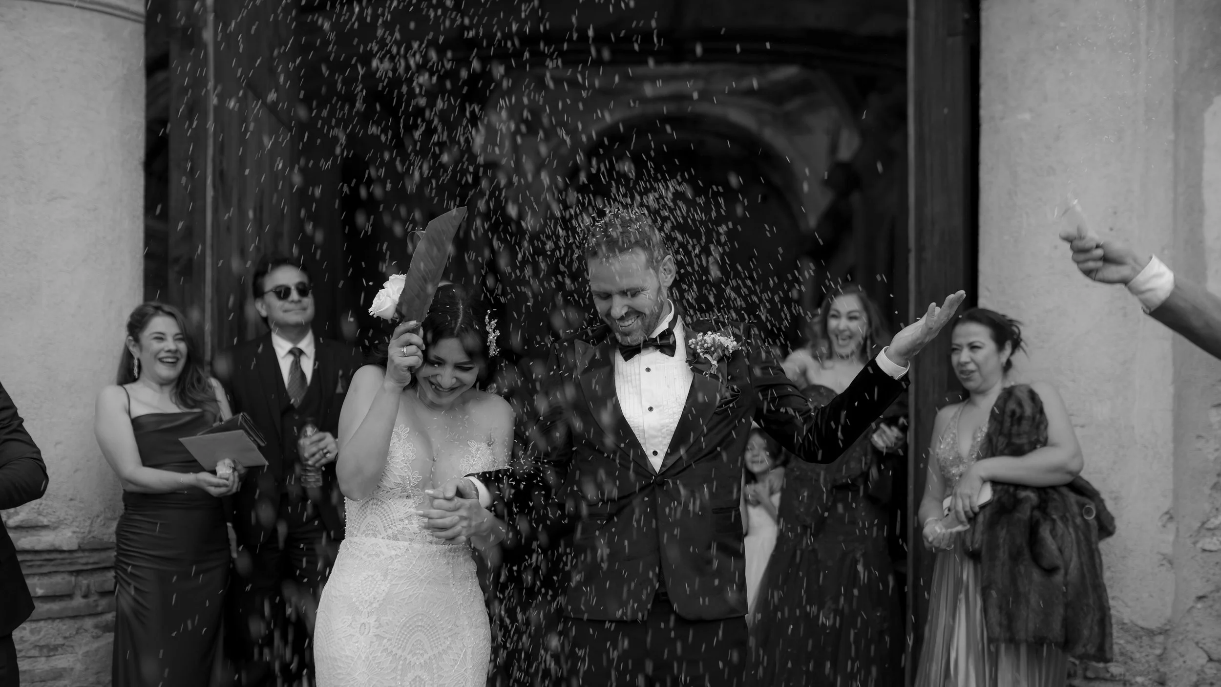Momentó de la boda con la pareja principal saliendo de la iglesia, rodeada de amigos lanzando arroz, en blanco y negro.