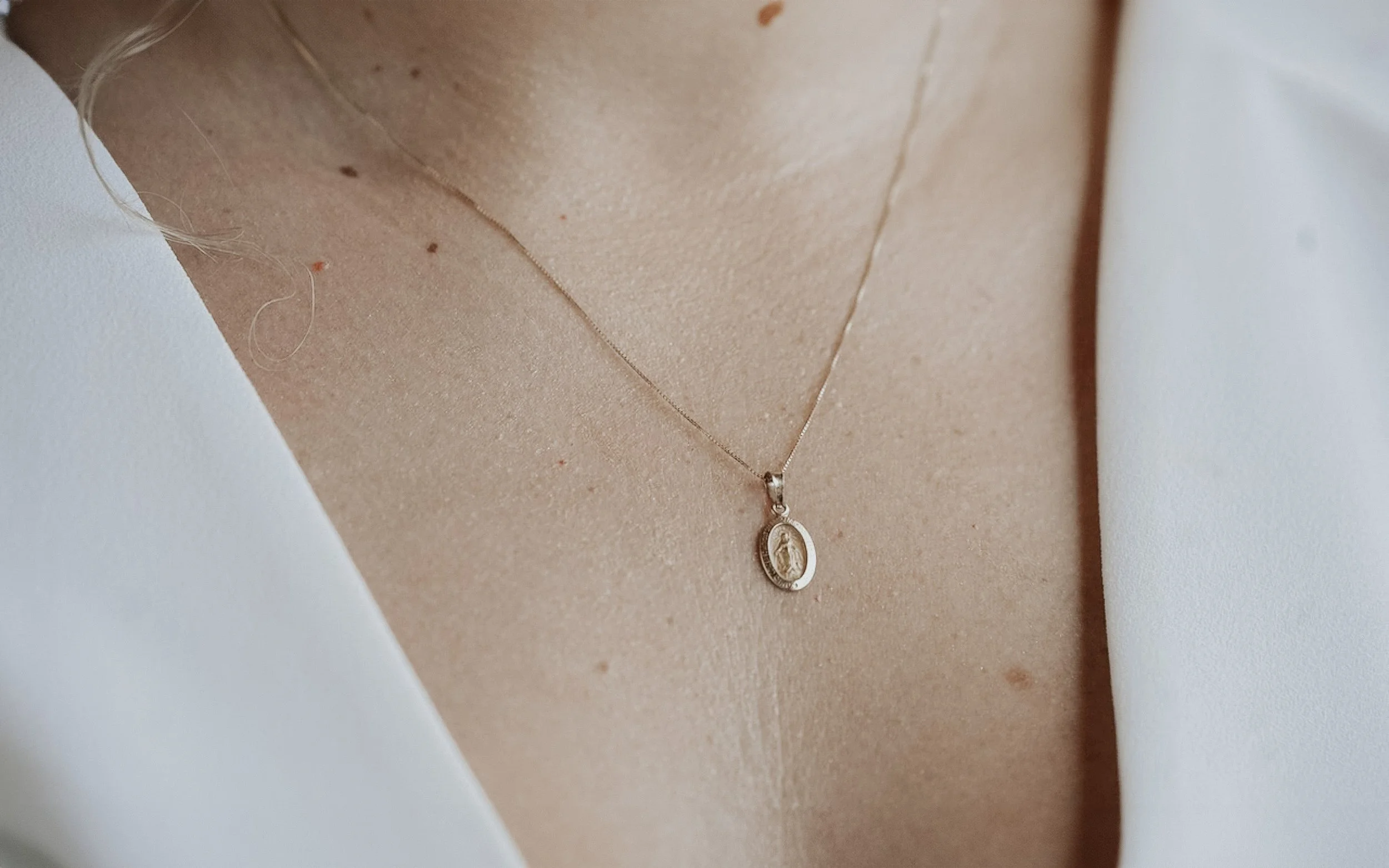 Cerca de la clavícula una cadena delgada con un colgante ovalado que tiene una figura en su interior, sobre piel desnuda y con ropa blanca a los lados.