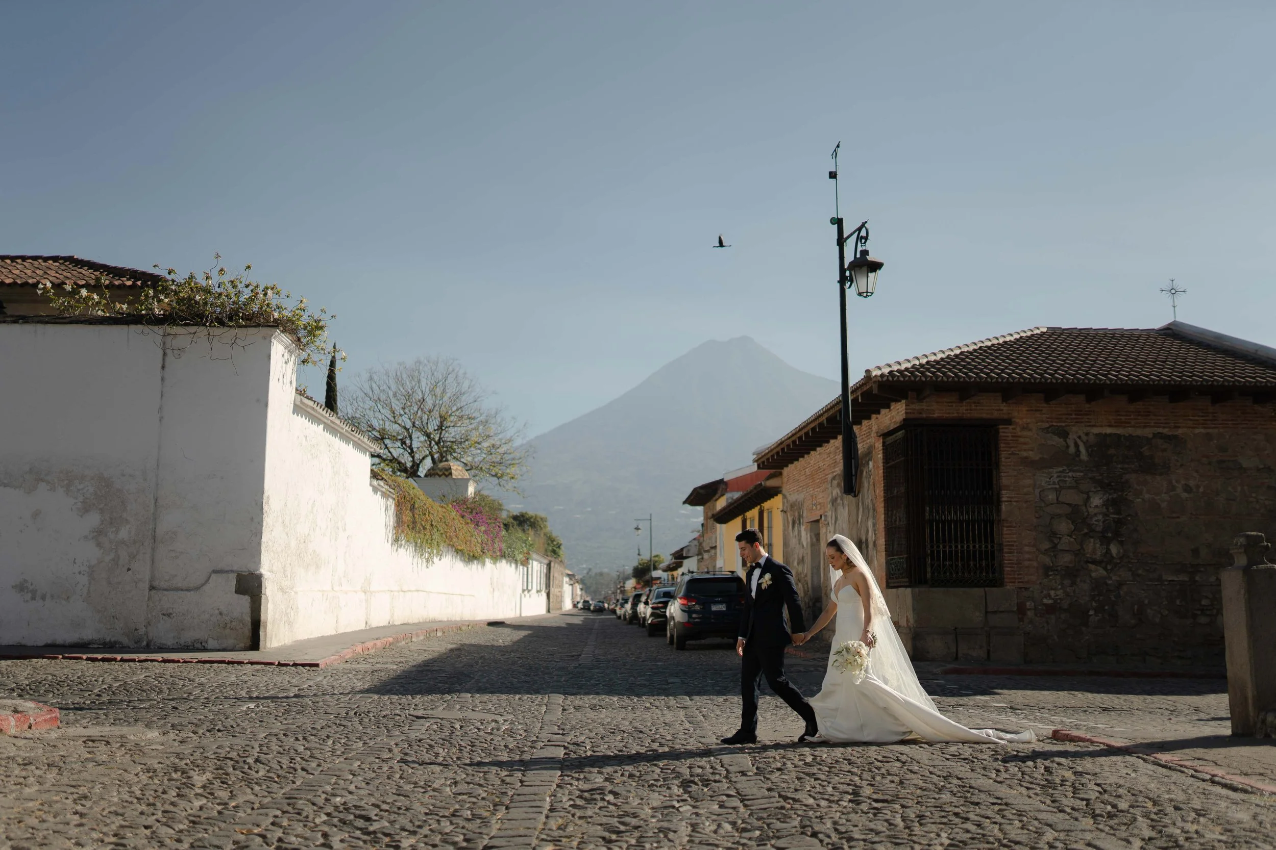 Una pareja de recién casados caminando de la mano en una calle empedrada, con una montaña en el fondo y cielo despejado.