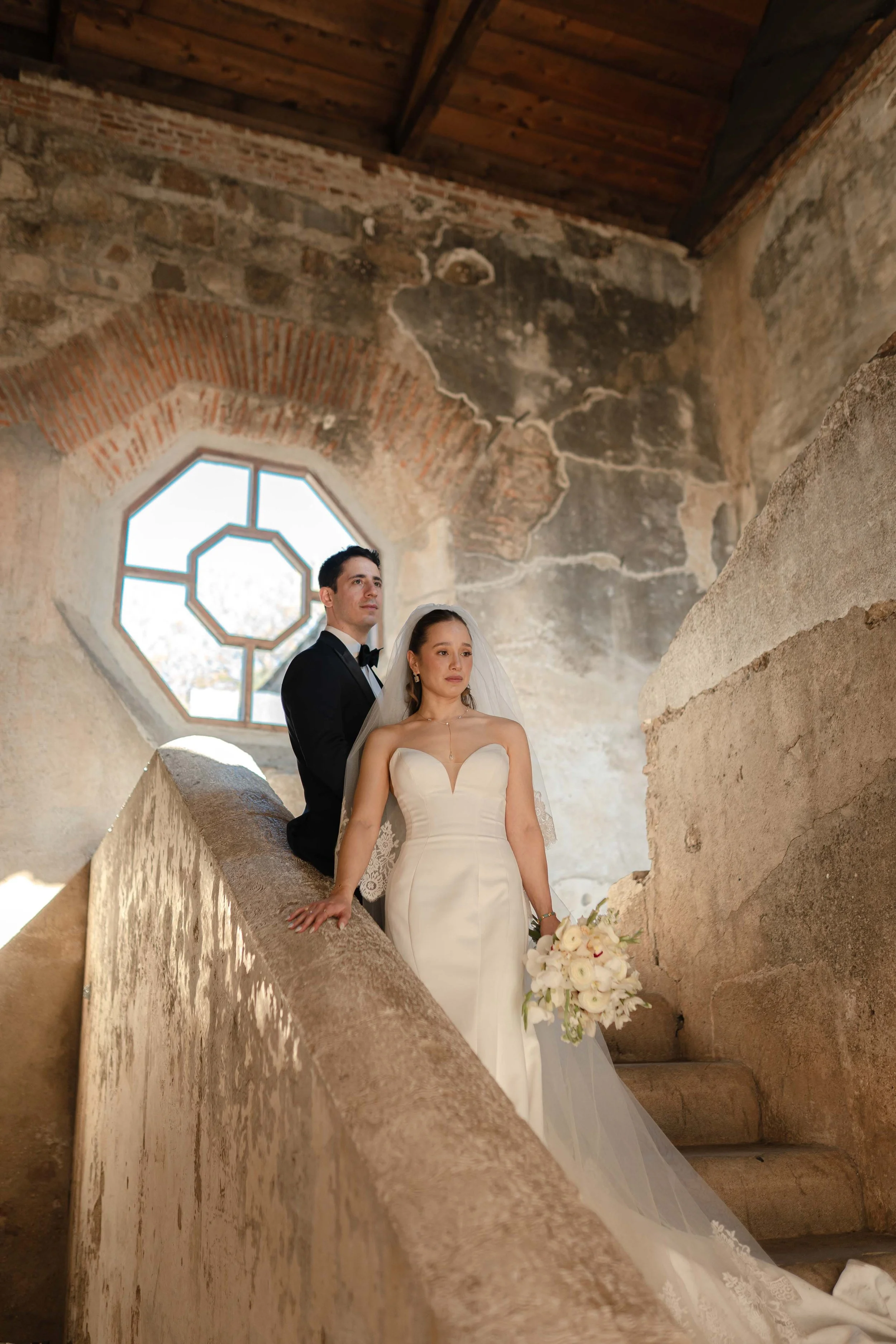 Pareja de novios en una antigua iglesia con paredes de piedra y una ventana decorativa detrás, la novia en vestido de novia y el novio en traje negro.