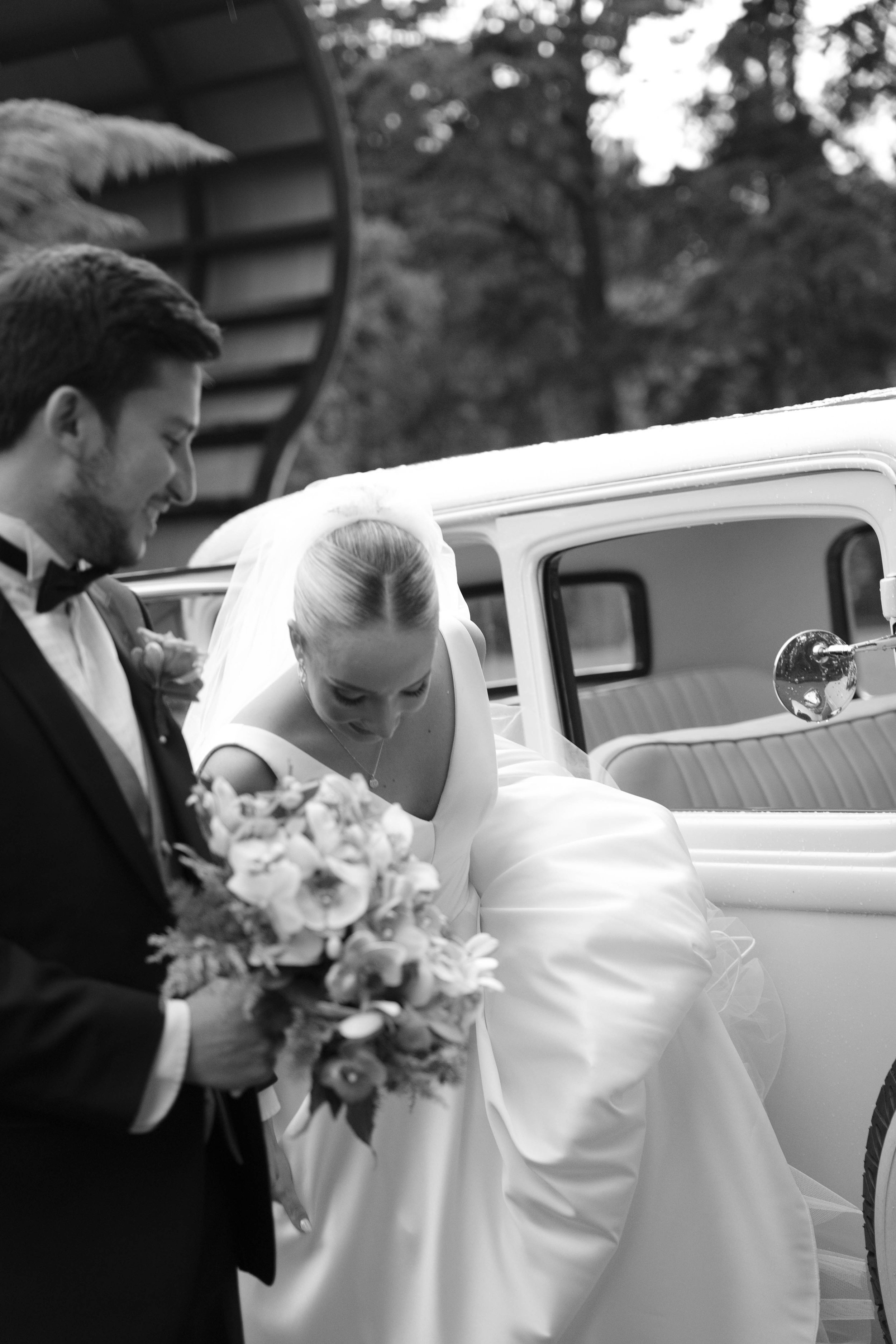 Pareja de recién casados en su boda, el novio en traje y la novia en vestido de novia, acercándose a un coche clásico blanco.