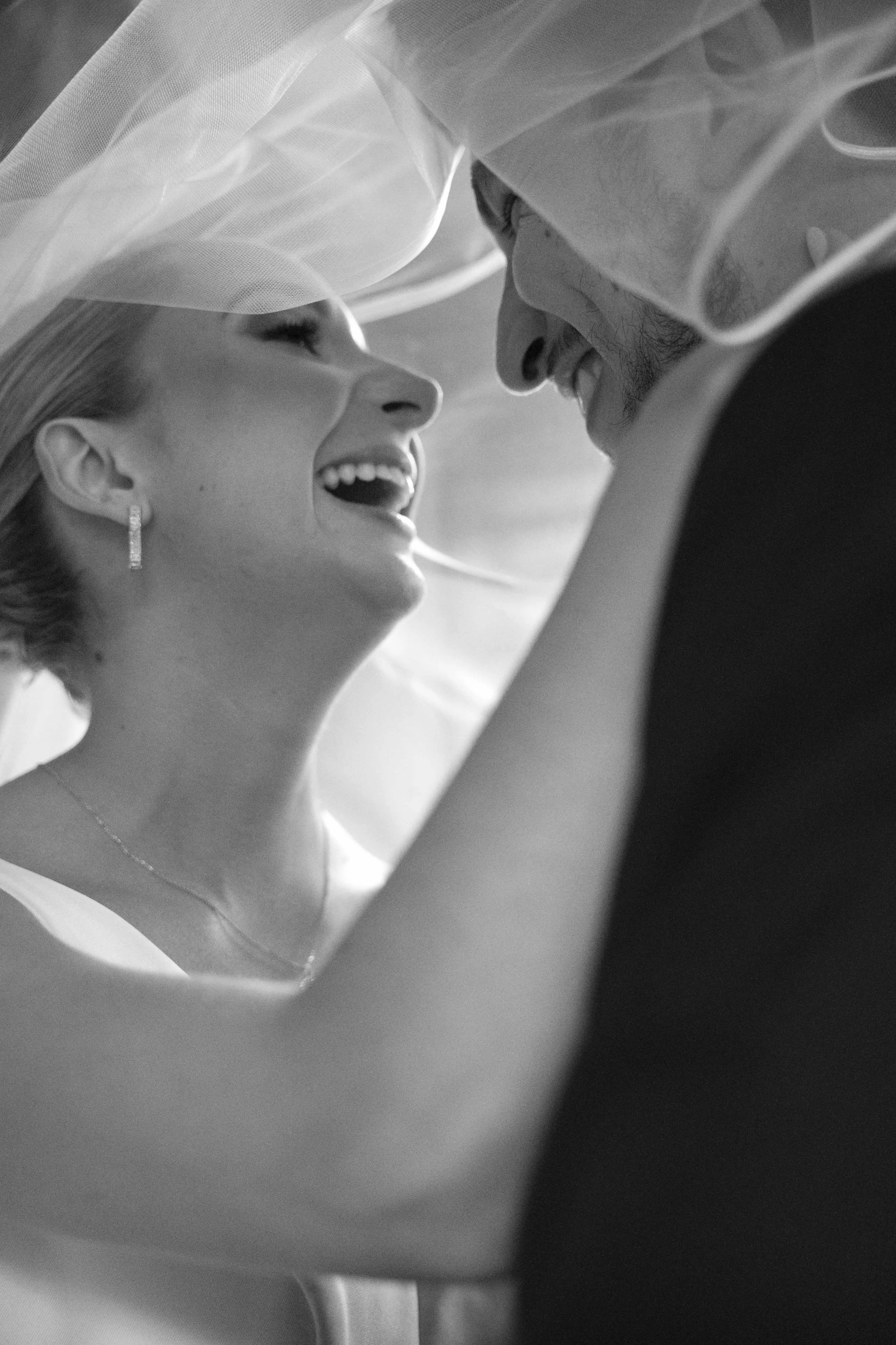 Una pareja de novios riendo en su boda, la novia con velo y el novio con barba, rodeados de tela y con expresión de felicidad.
