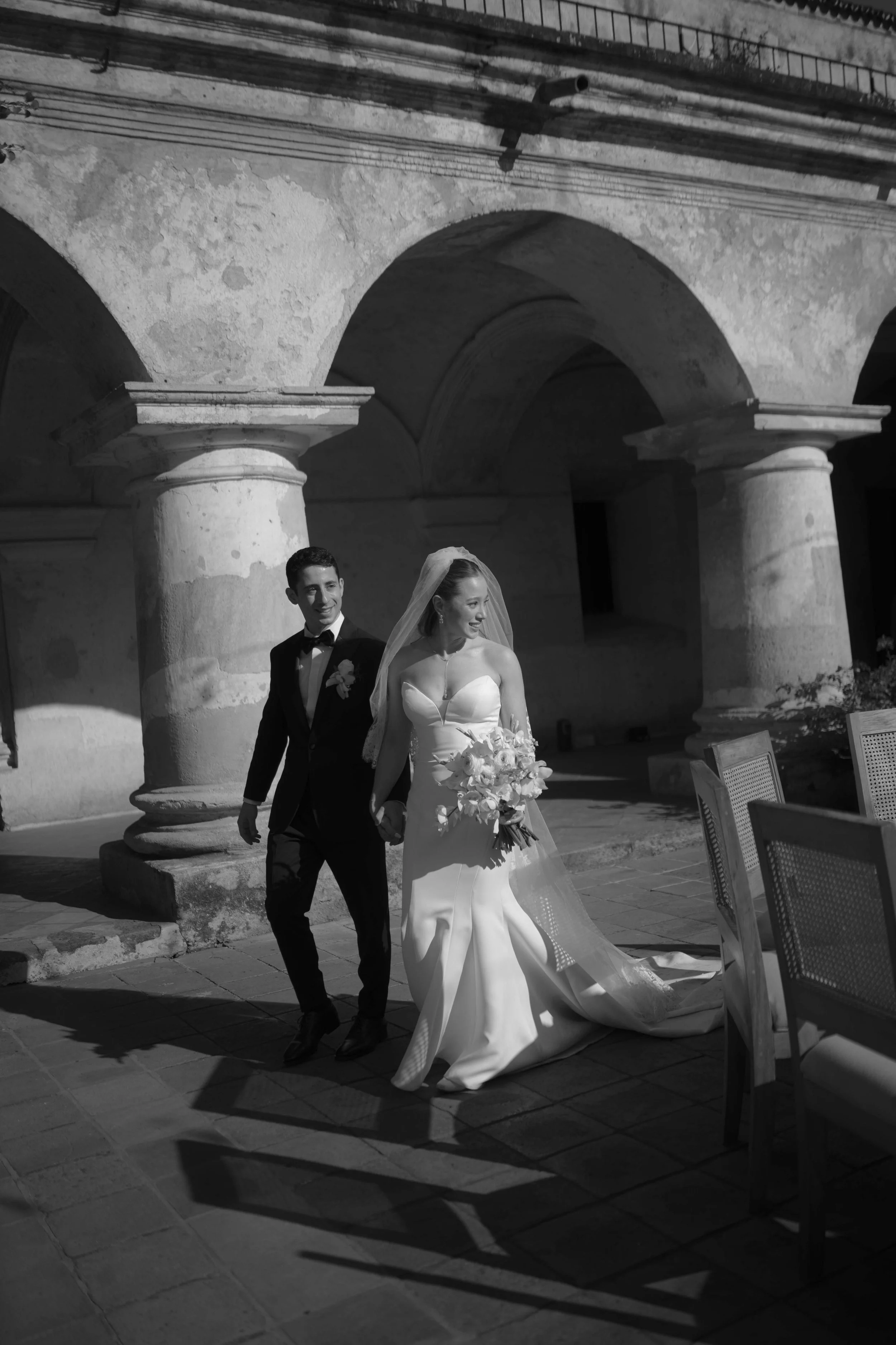 Pareja de novios de bodas caminando en un lugar con columnas y arcos, vista en blanco y negro.