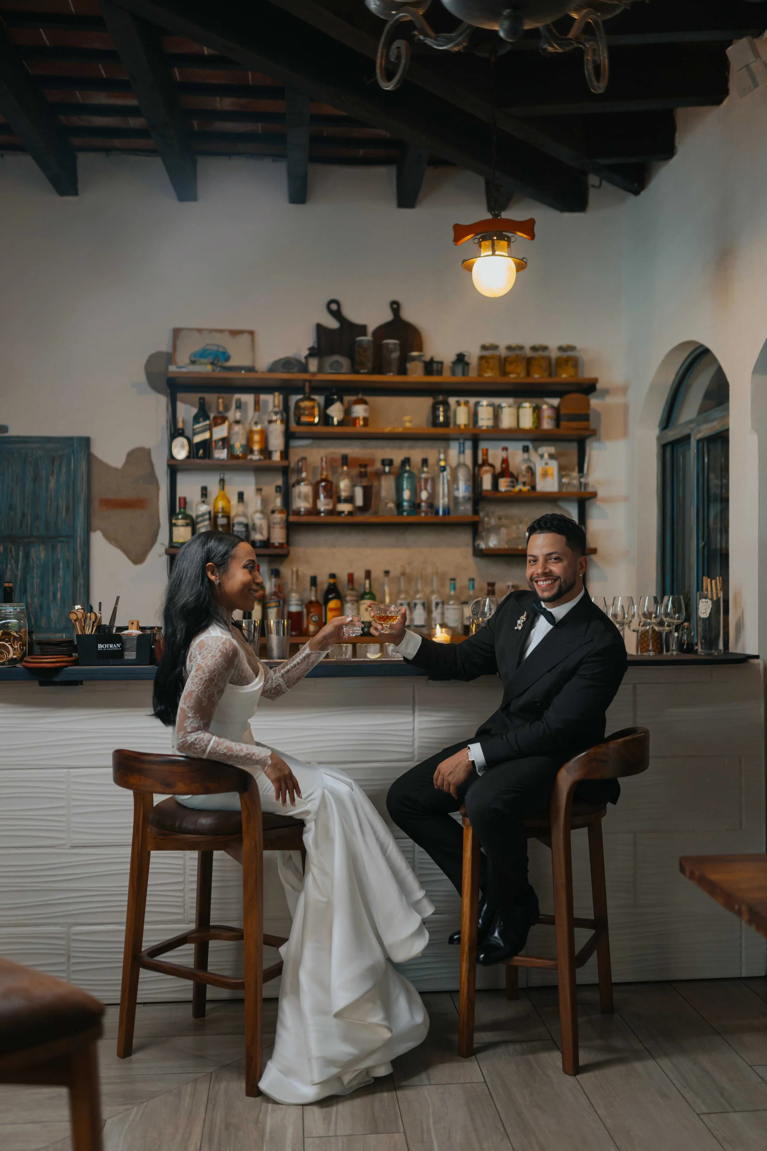 Pareja de novios brindando en un bar, ella con vestido de novia y él en tuxedo, sonriendo.
