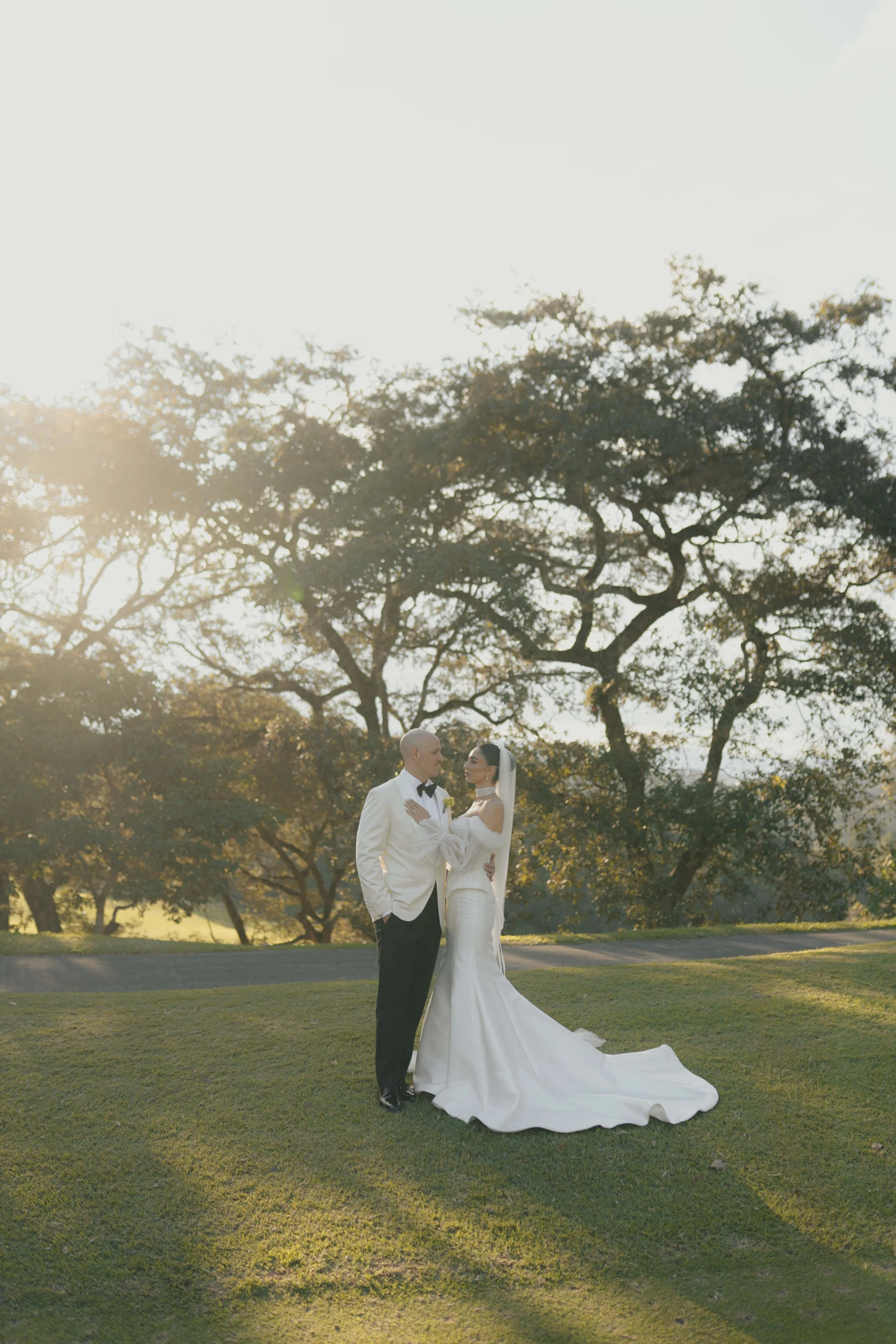 Pareja de novios en vestido de boda y esmoquin en un parque, con árboles de fondo y luz cálida de atardecer.