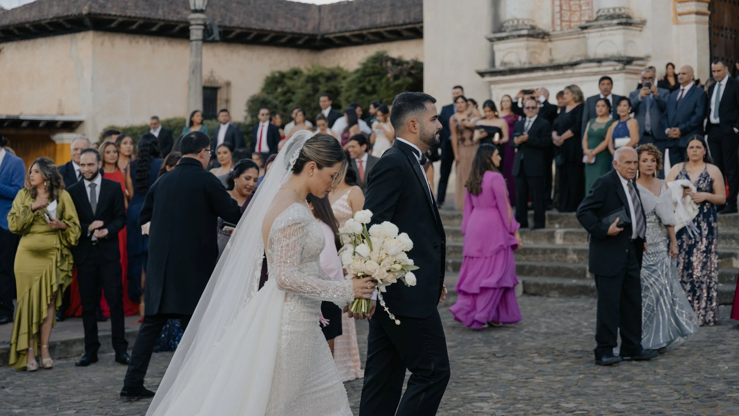 Una pareja de novios en una boda, caminando frente a una multitud de invitados en una iglesia antigua, en una calle empedrada. La novia lleva vestido blanco y sostiene un ramo de flores blancas, mientras que el novio viste traje negro, rodeados de in