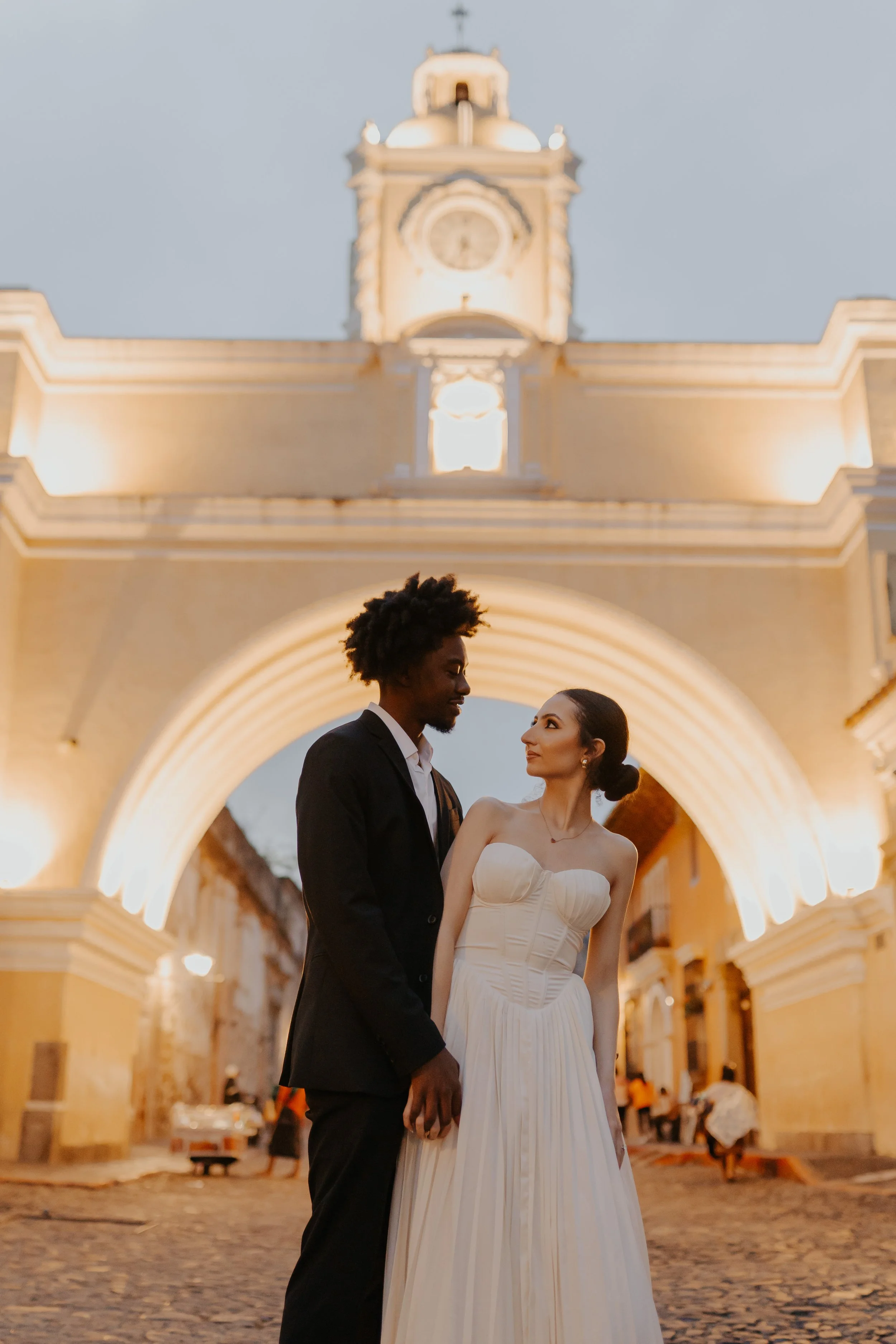 Una pareja vestida de boda, caminando en una calle empedrada frente a un arco iluminado y un reloj en una torre antigua, durante la noche.