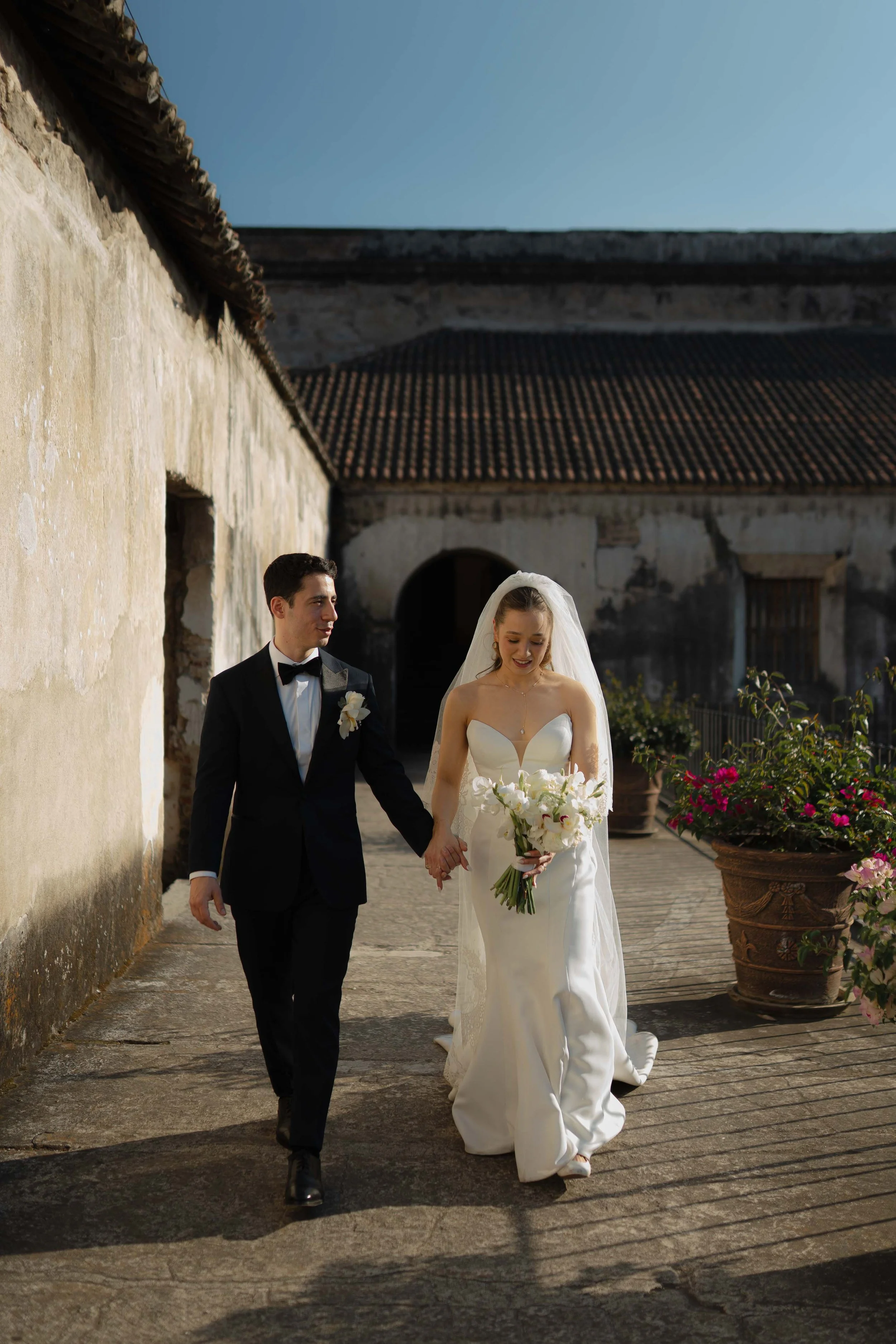 Una pareja de recién casados caminando juntos en un patio, ella con vestido de novia y él con traje negro, sosteniendo sus manos y rodeados de macetas con flores.