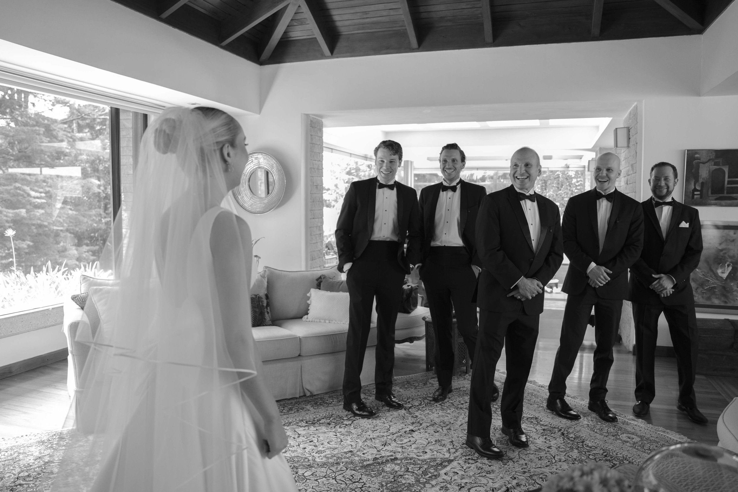 Una novia con vestido y velo mira a un grupo de cinco hombres en trajes formales con pajarita, en una sala bien iluminada.