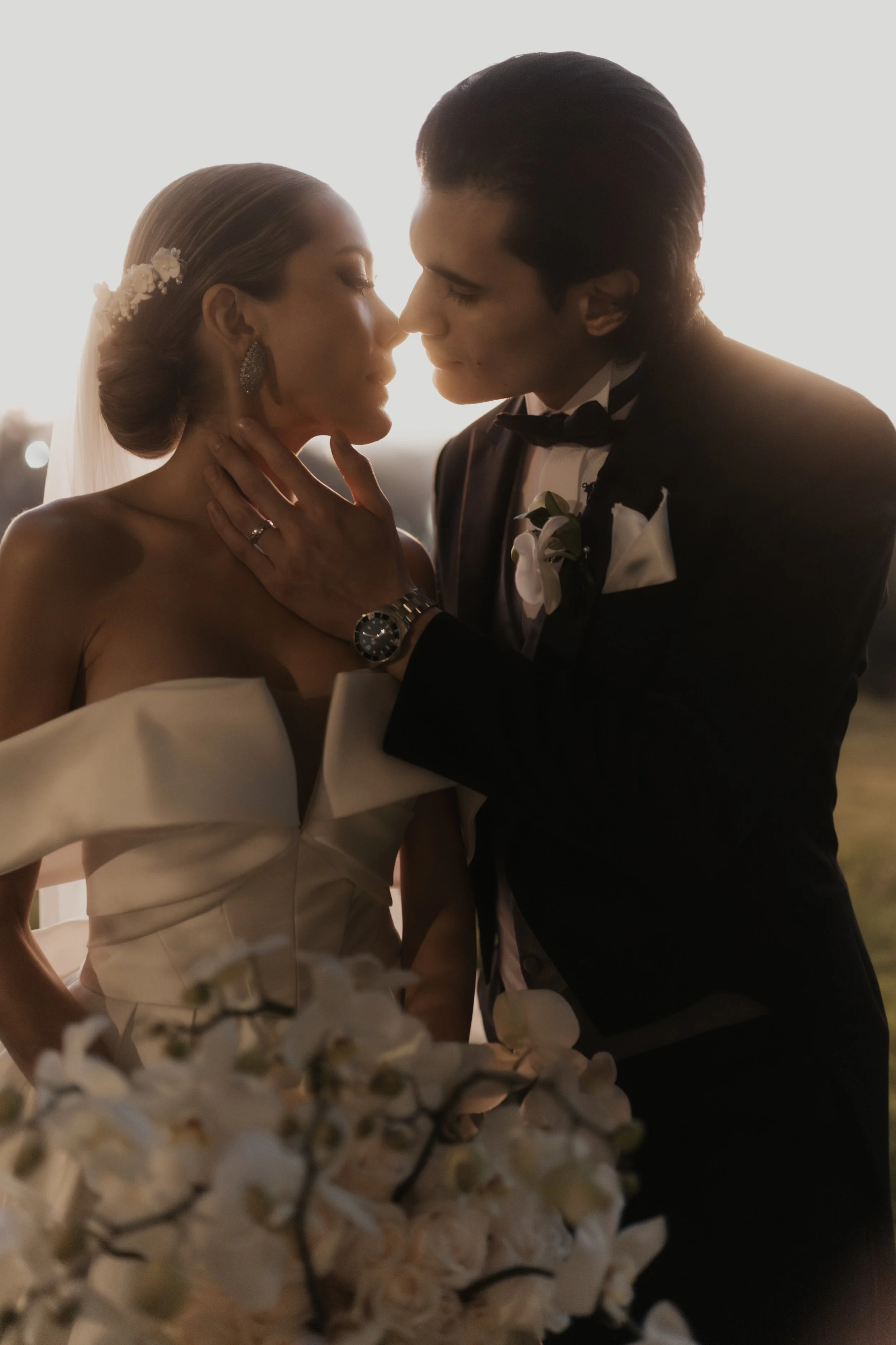 Pareja de novios en rito de boda, con la mujer vestida de novia y el hombre de esmoquin, en un momento íntimo con fondo difuso y flores blancas en primer plano.