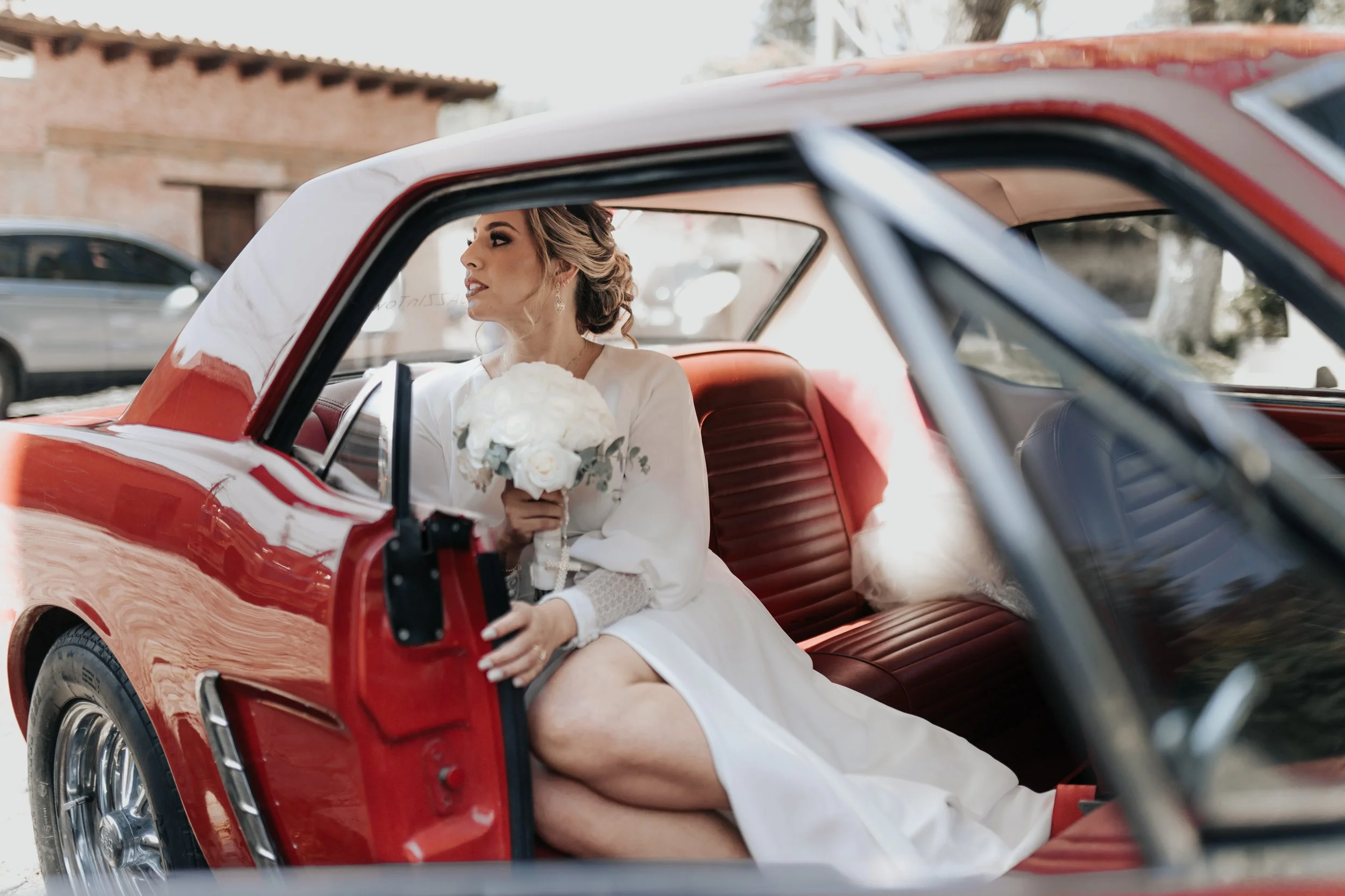 Mujer con vestido de novia sentado en interior de automóvil rojo clásico, sosteniendo un ramo de flores blancas.