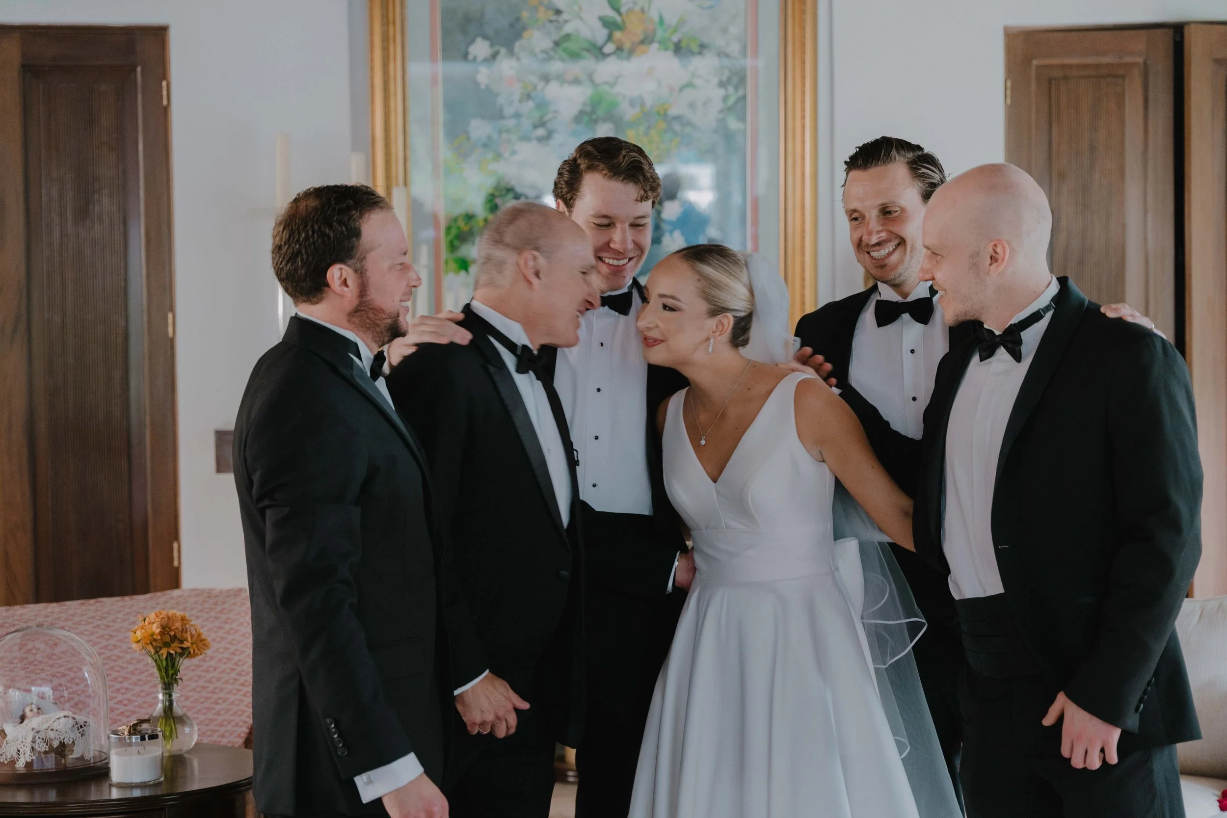 Grupo de siete personas en un momento de celebración en una boda, con la novia en el centro y rodeada de los otros seis hombres vestidos con esmoking, en un interior decorado con madera y arte en la pared