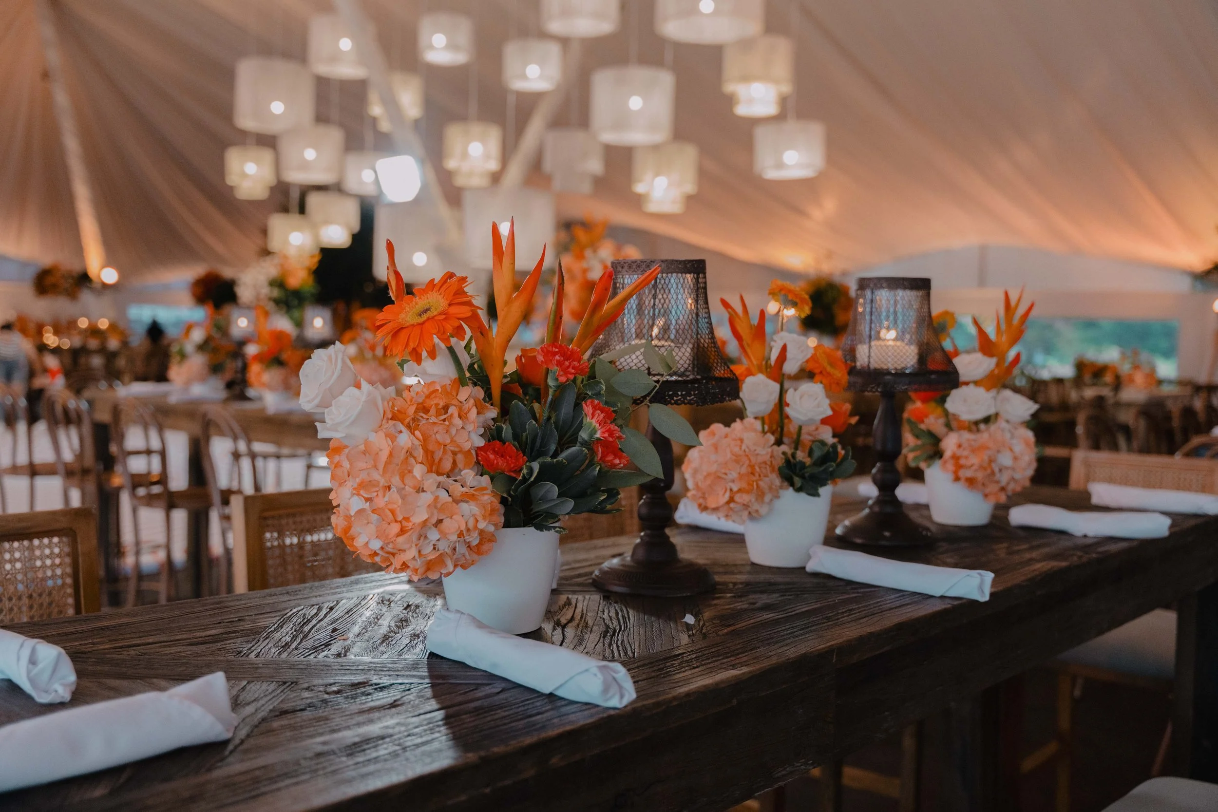 Decoración de mesa con arreglos florales en tonos naranjas y rosas, junto a candelabros negros, en un evento bajo una carpa con iluminación cálida y mesas largas.