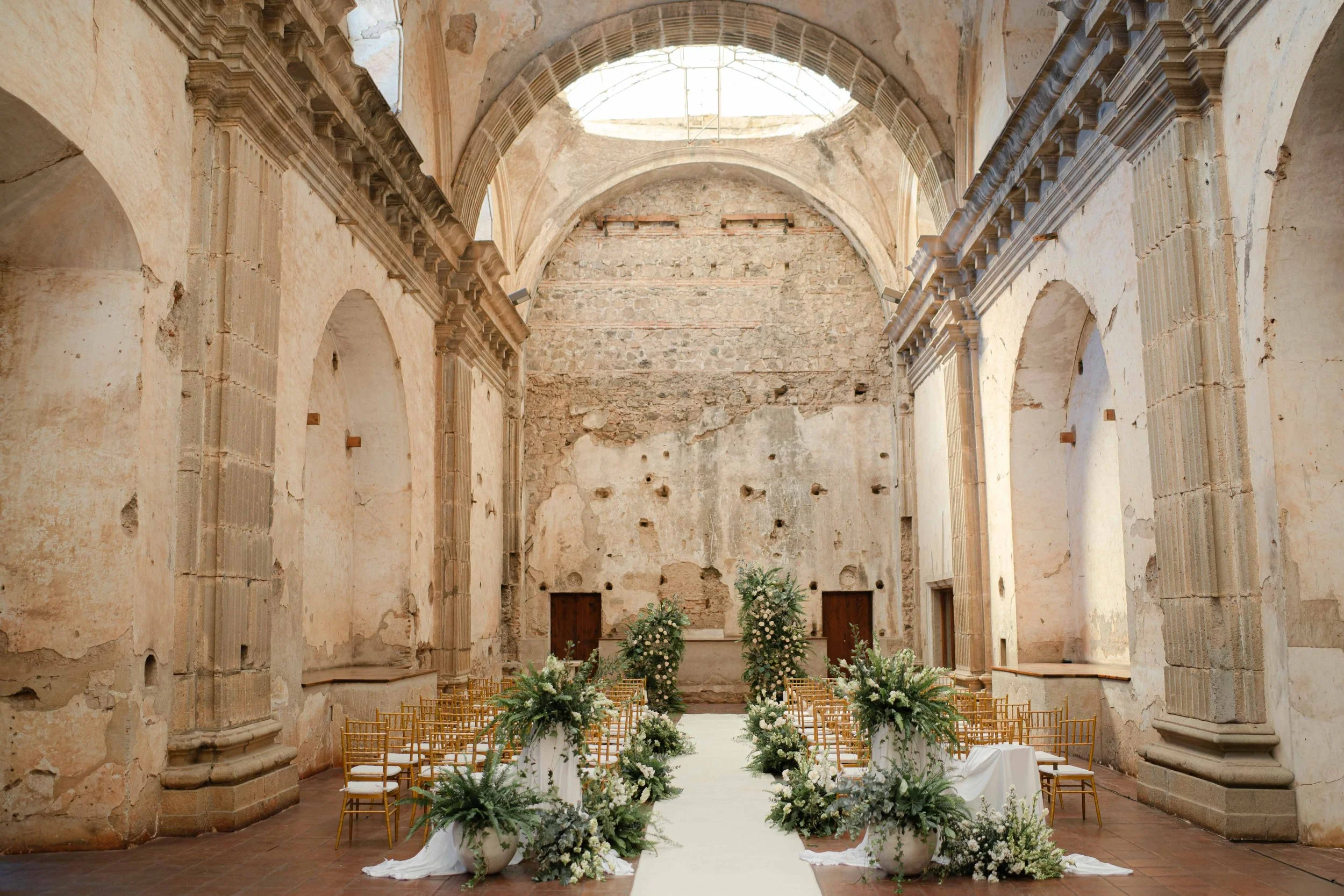 Interior de una iglesia antigua decorada para una boda, con sillas doradas, arreglos florales en jarrones y un pasillo en el centro.