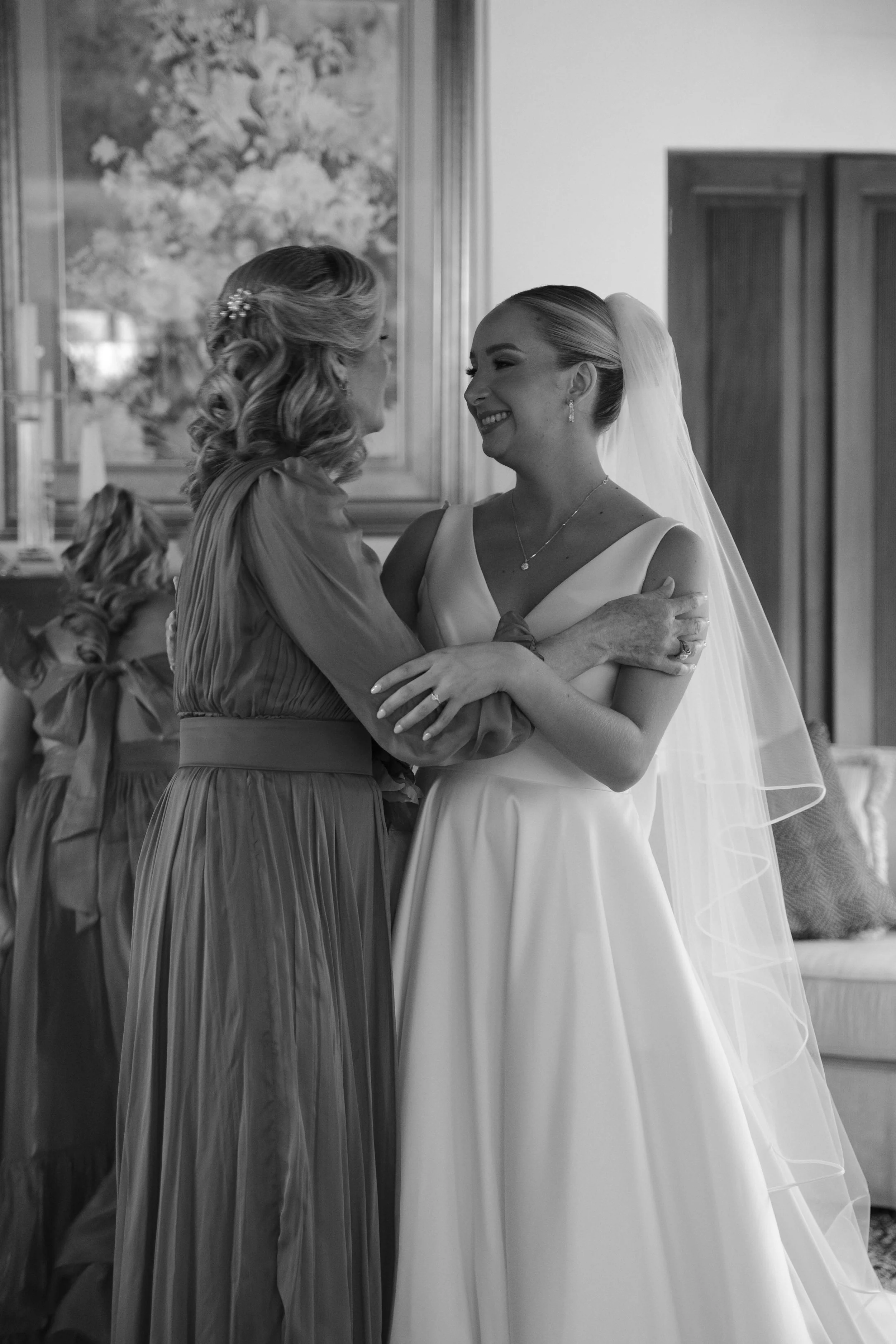 Mujer con vestido de novia sonriendo y abrazando a otra mujer en una ceremonia de boda, en un entorno interior.