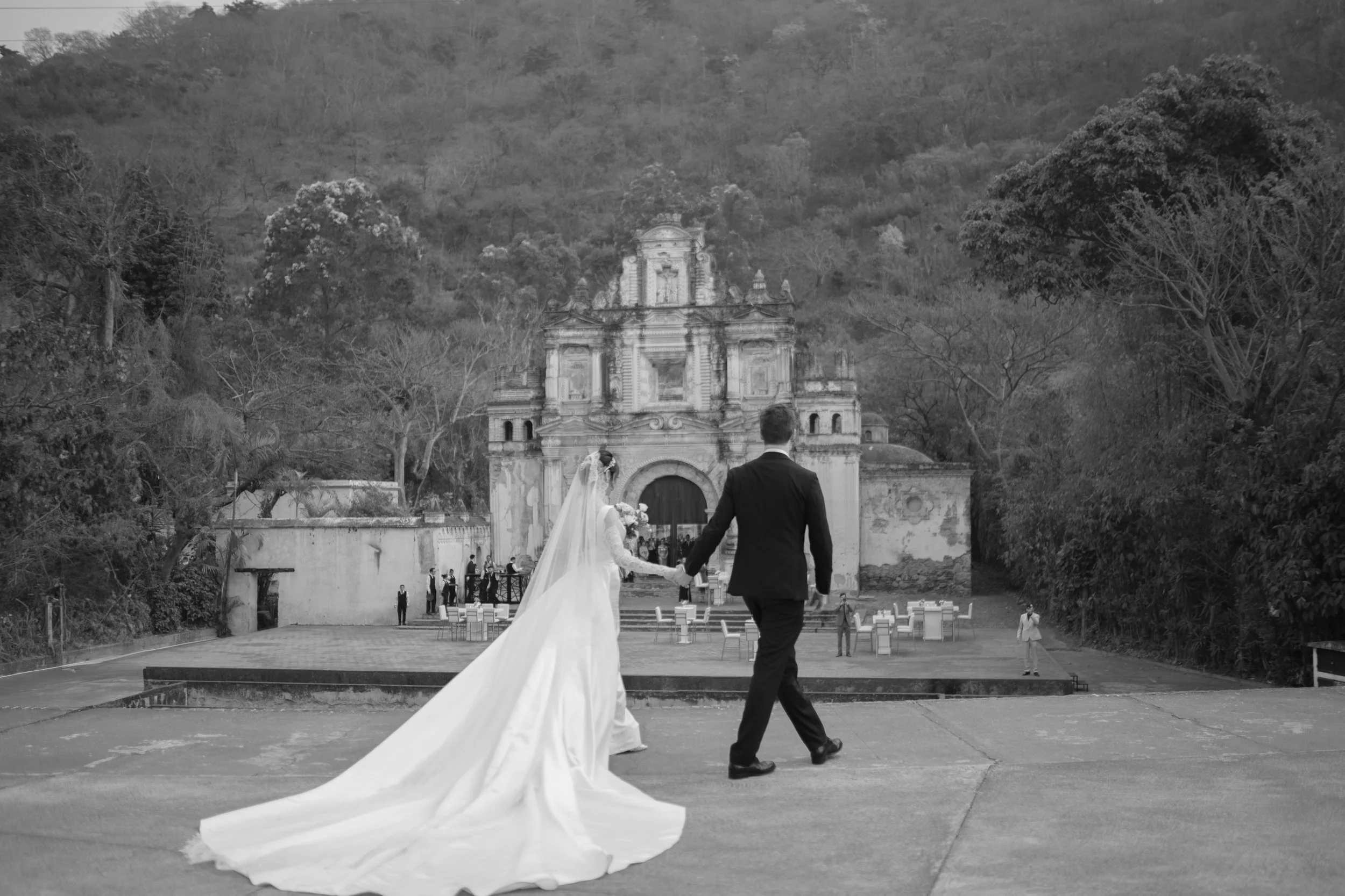 Pareja de novios caminando hacia una iglesia en  Antigua Guatemala en un entorno natural. La novia lleva vestido largo y velo, el novio lleva traje negro.