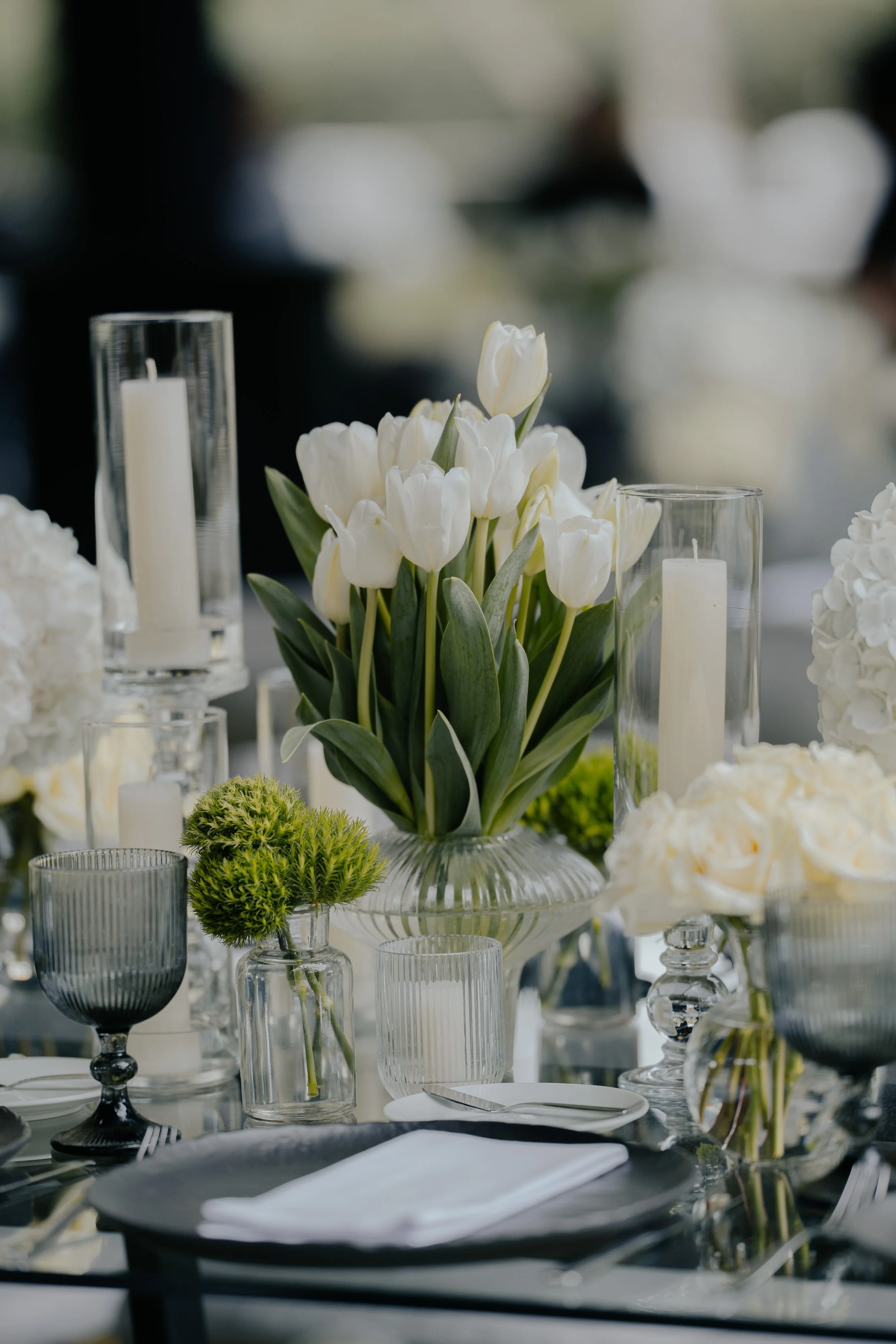 Mesa decorada con centros de flores blancas, velas en candelabros de cristal y vajilla elegante en tonos oscuros.