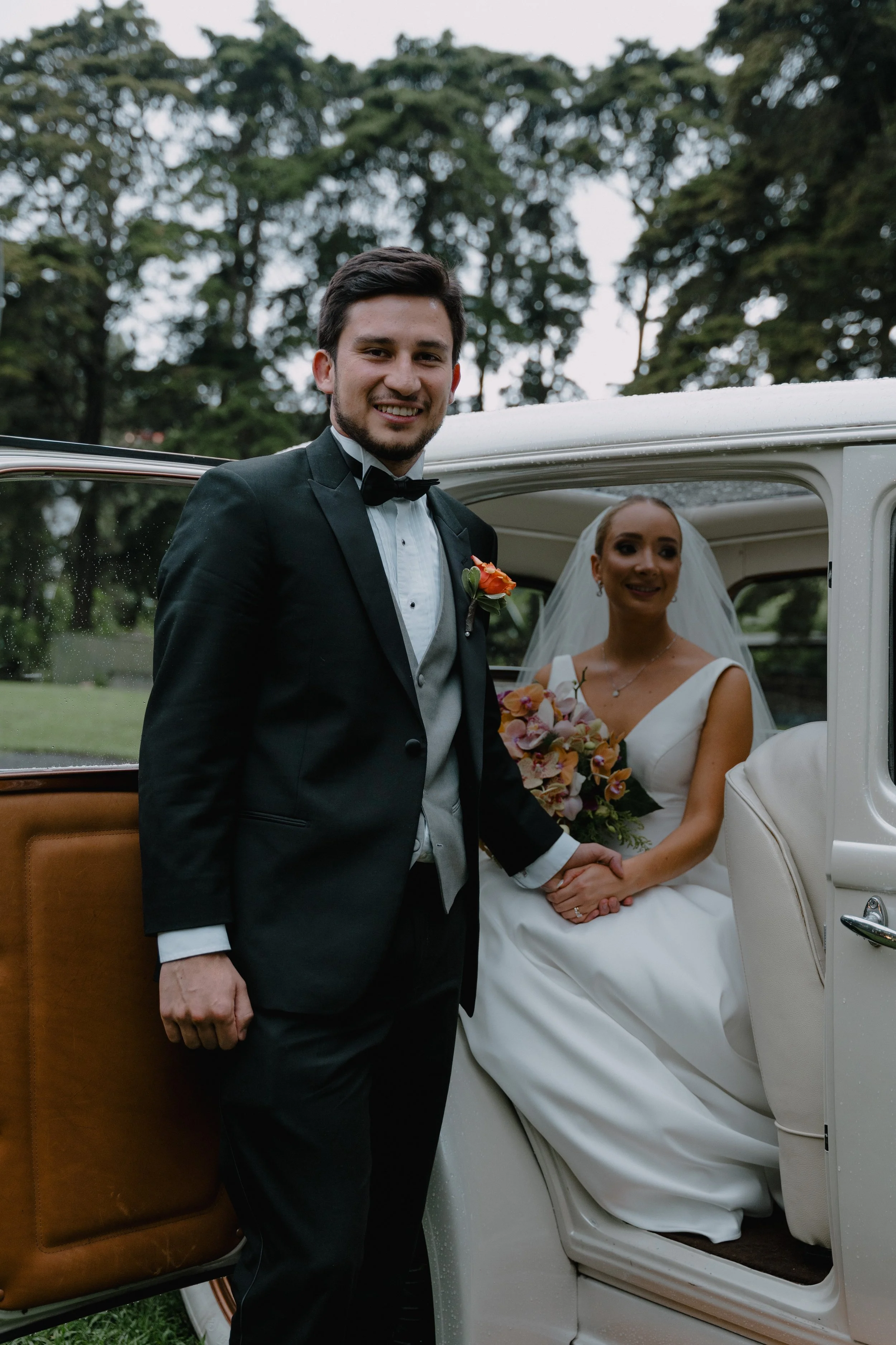 Una pareja de recién casados posando junto a un automóvil clásico al aire libre, ella con un vestido de novia y él con un esmoquin, en un entorno natural con árboles de fondo.