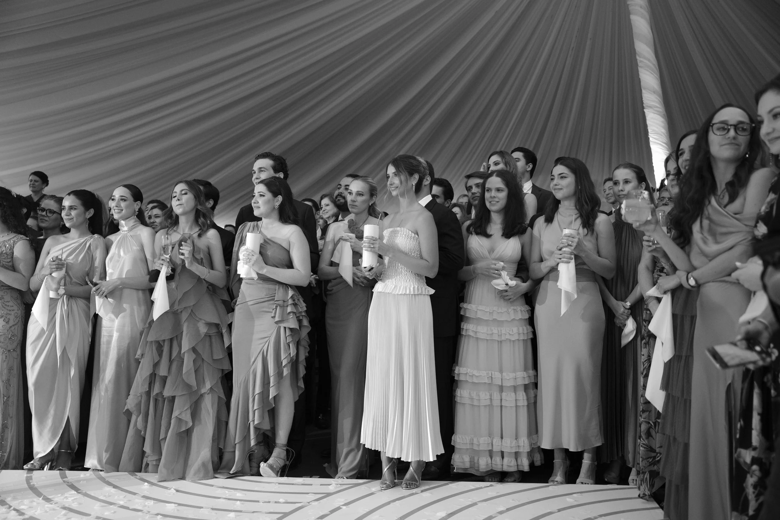 Grupo de personas vestidas con ropa formal en un evento, algunas sosteniendo velas y bebidas en un ambiente decorado con tela en el techo.