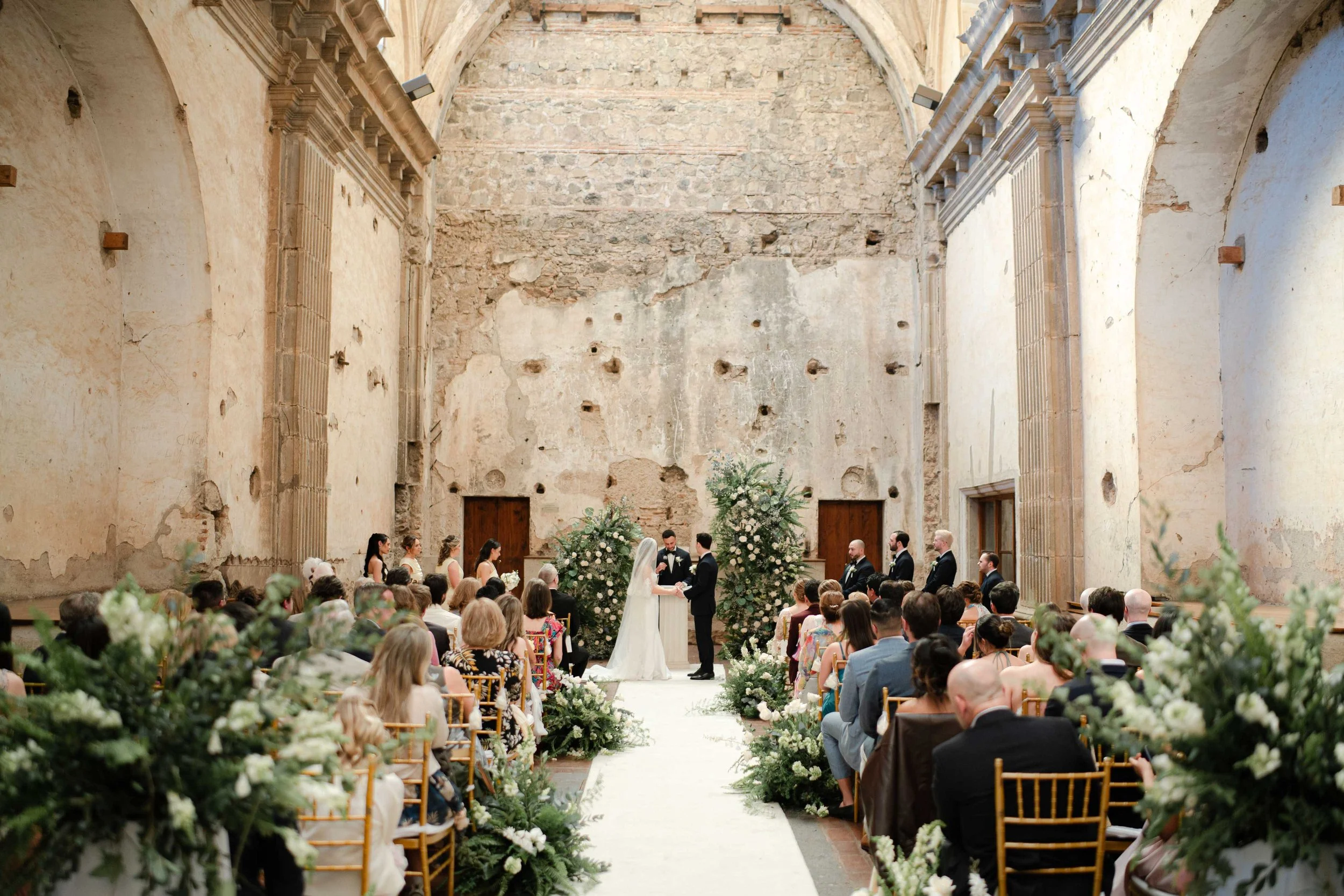 Una ceremonia de boda en una iglesia antigua con paredes de piedra, decorada con muchas flores blancas y verdes, con pareja casándose frente a una congregación.