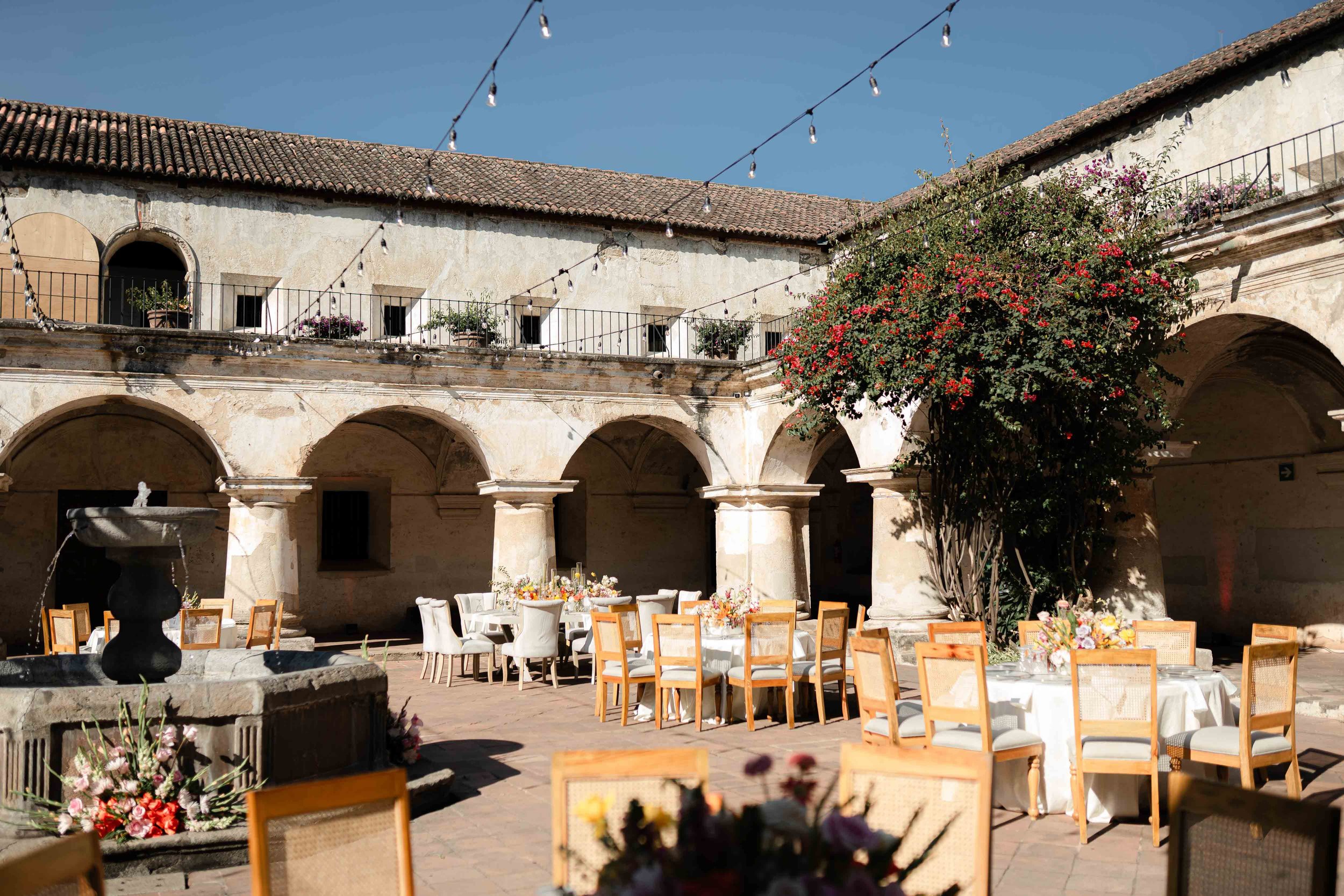 Decoración de evento en un patio con mesas y sillas blancas, fuente y árbol con flores rojas.
