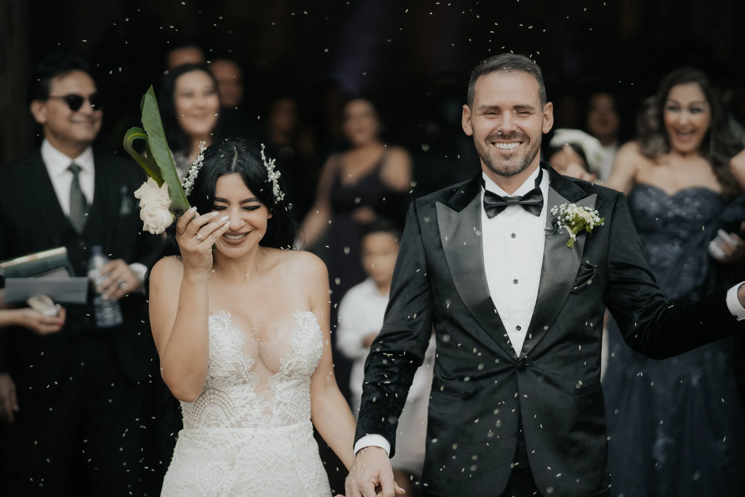 Pareja de recién casados caminando felizmente entre amigos y familiares, lanzando confeti en una celebración nupcial, ella con vestido de novia y él con tuxedo, en un ambiente alegre y festivo.