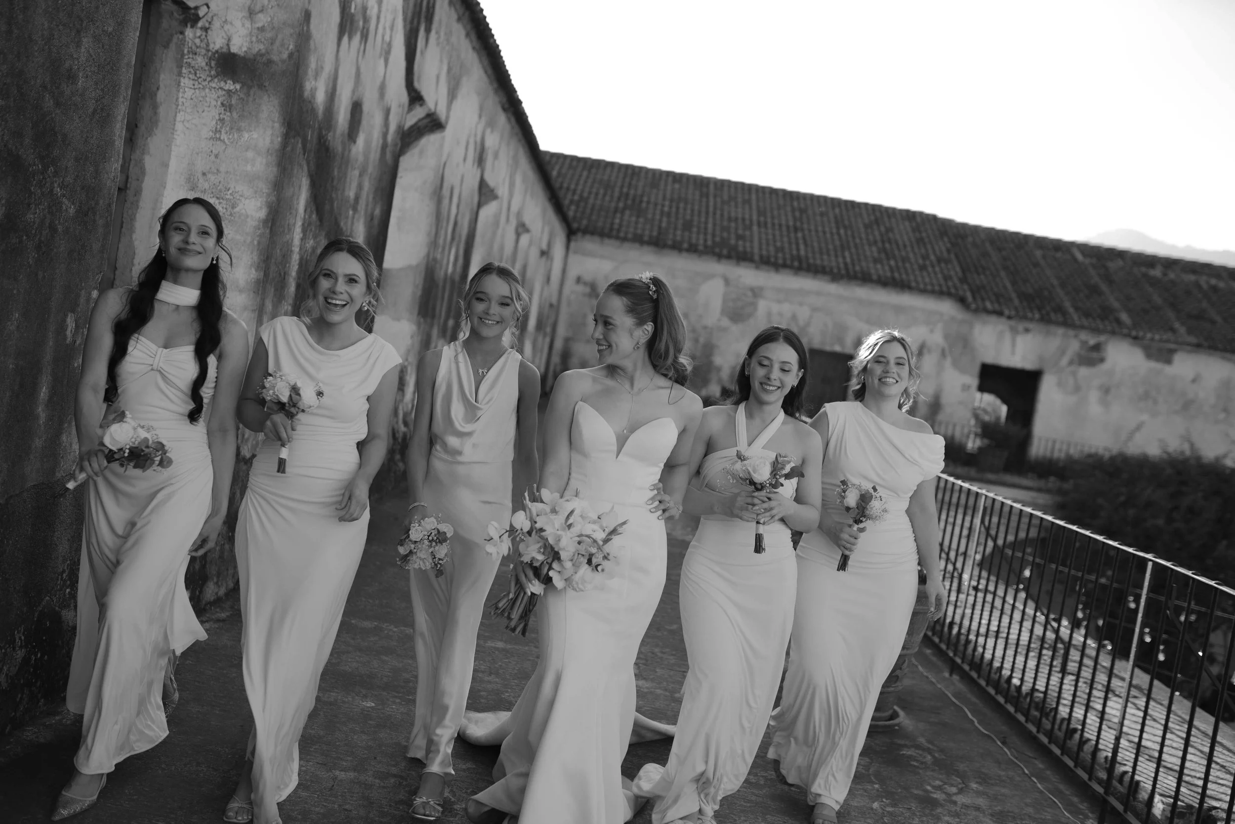 Grupo de siete mujeres en vestidos de novia y damas de honor caminando juntas en un pasillo al aire libre, sonriendo y mostrando sus ramos de flores, en un entorno histórico con paredes de piedra.