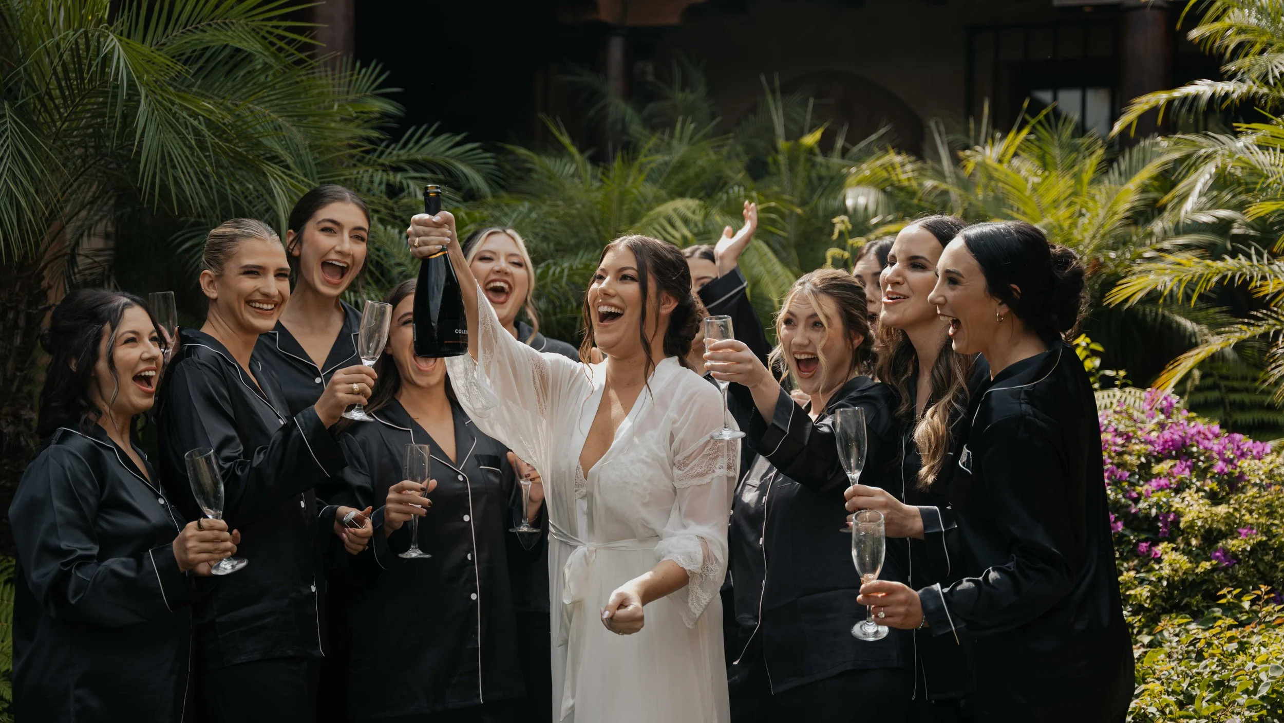Grupo de mujeres celebrando con champán en un jardín, algunas con pijamas negras y una con vestido blanco. La mujer con vestido blanco está levantando una botella de champán, todas sonrientes y alegres.