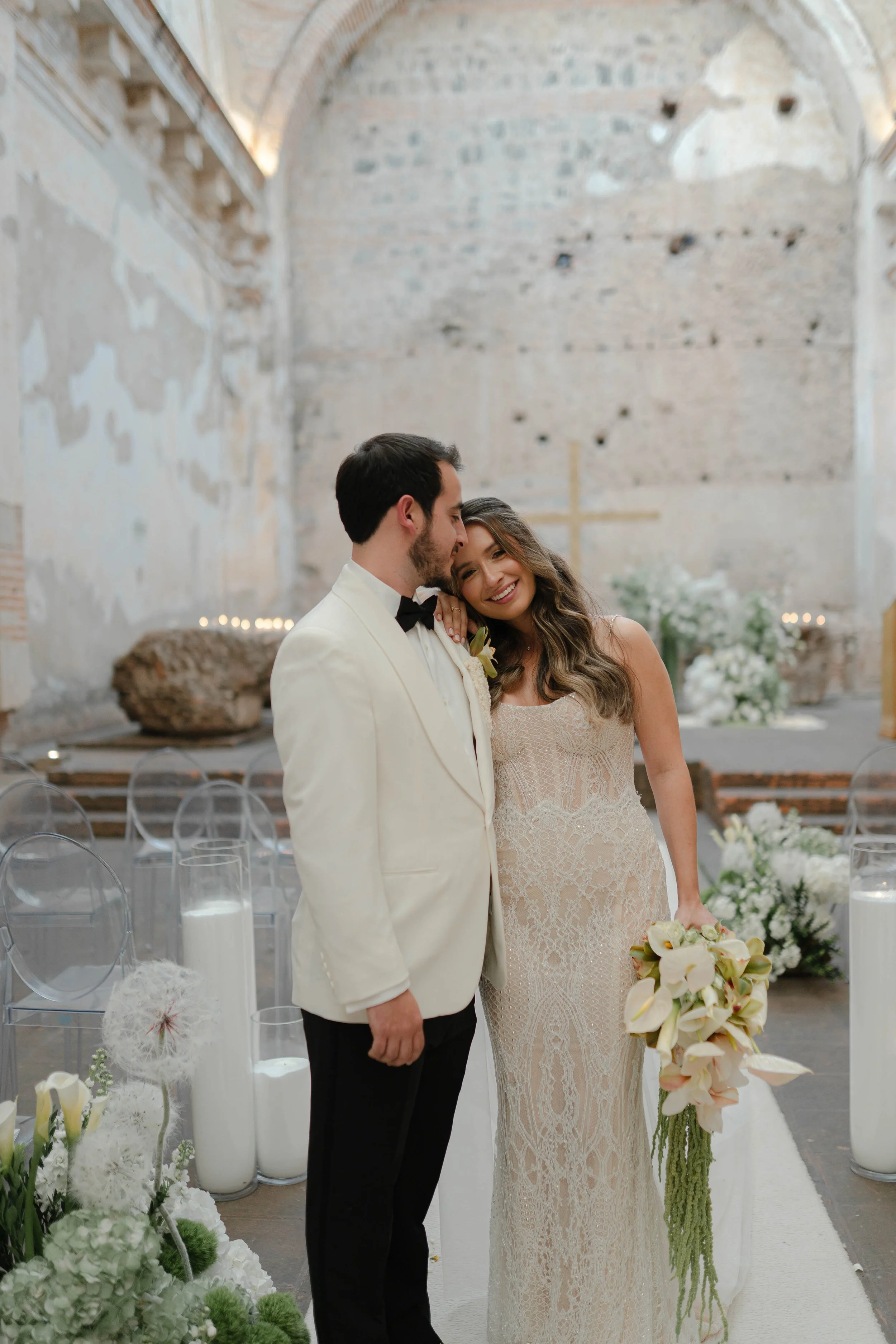 Una pareja de boda dentro de la iglesia, con el novio vestido con esmoquin blanco con pajarita negra y la novia con vestido de encaje y bouquet de flores, sonriendo y compartiendo un momento de felicidad.