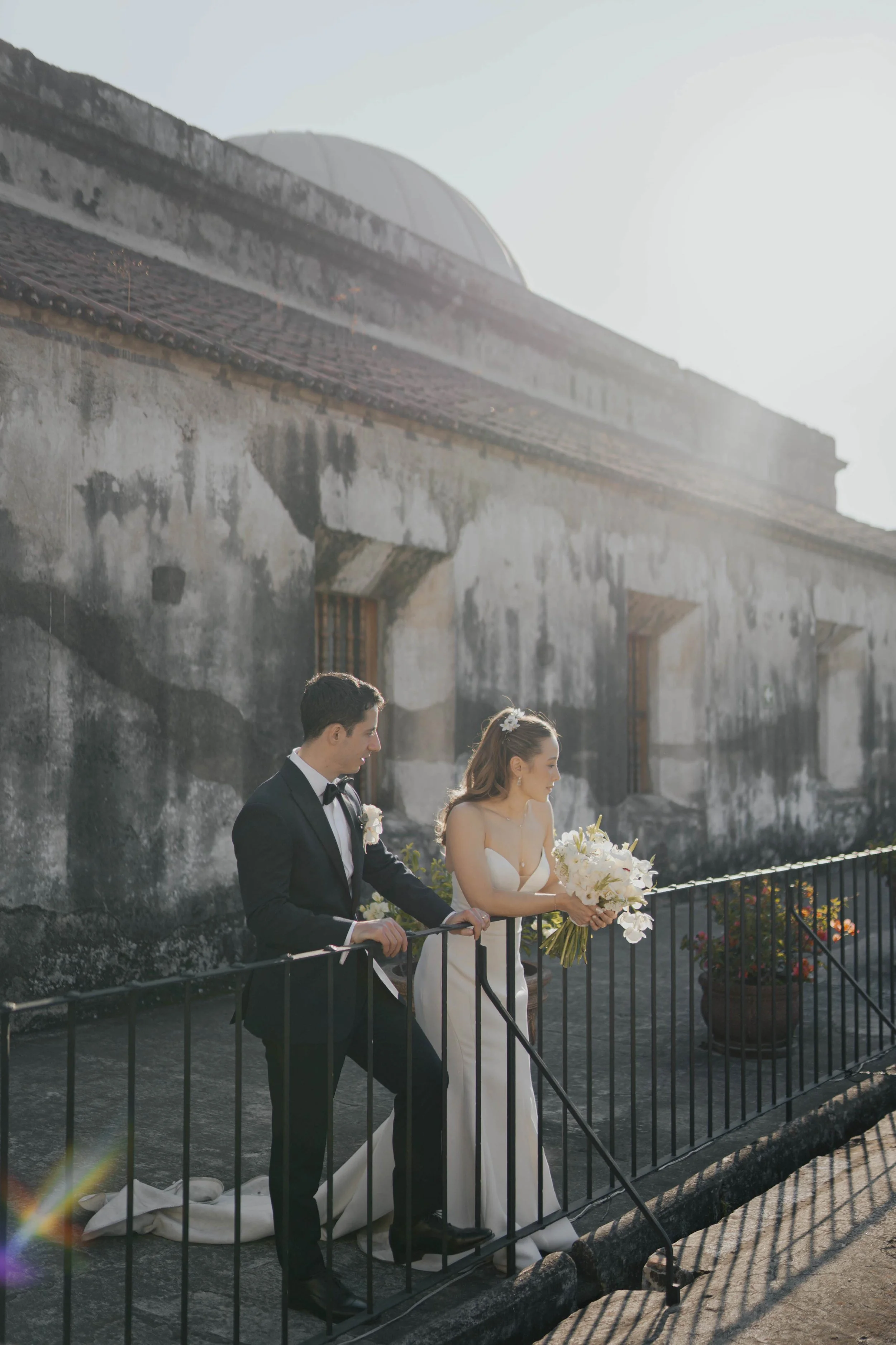Pareja de recién casados en un balcón, la novia con vestido blanco y ramo de flores, el novio con esmoquin, en un ambiente al aire libre con muro envejecido y plantas en macetas.