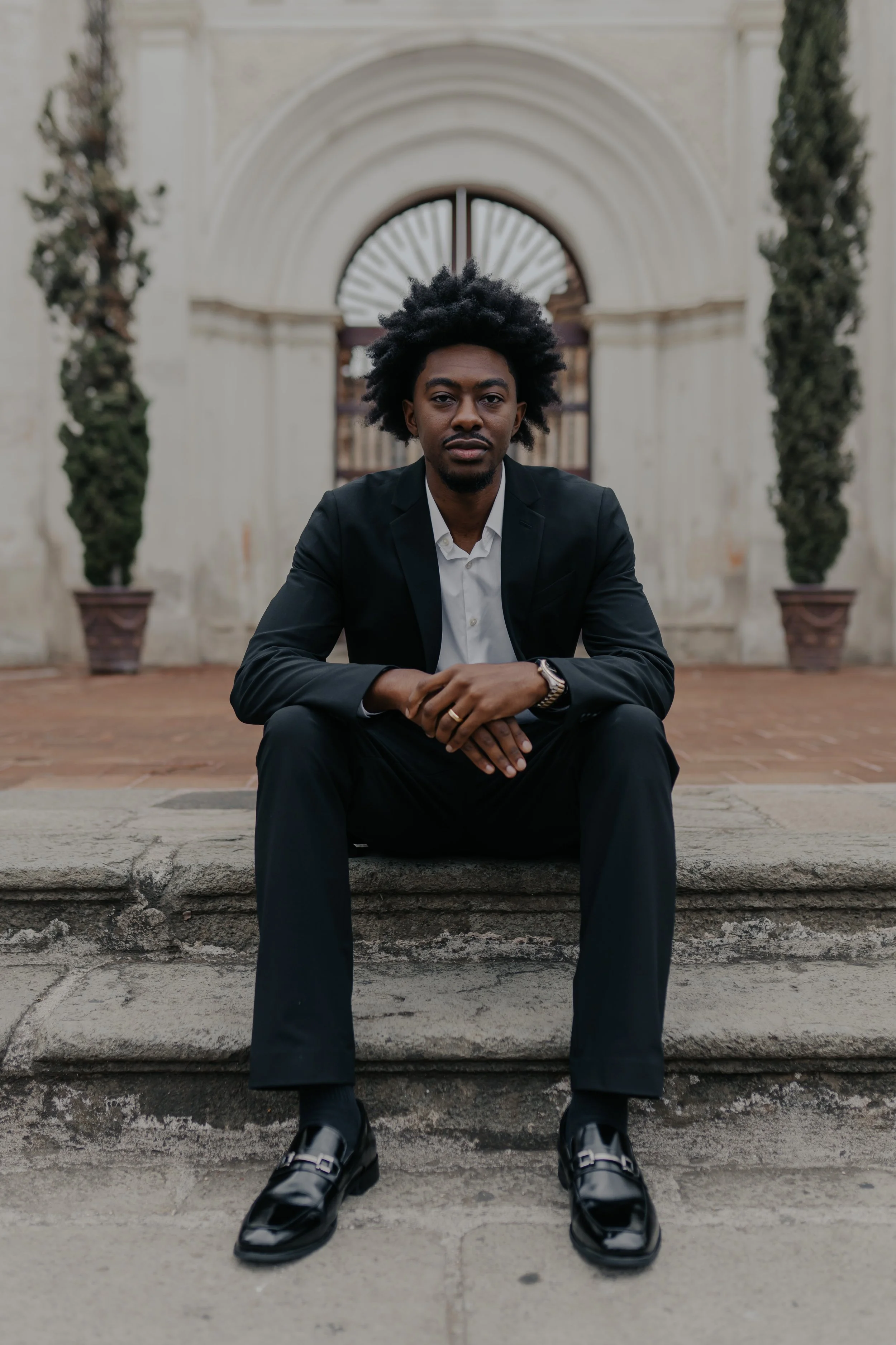Joven hombre con traje negro sentado en las escaleras frente a una estructura histórica de piedra con arcos, con dos árboles en macetas a cada lado.