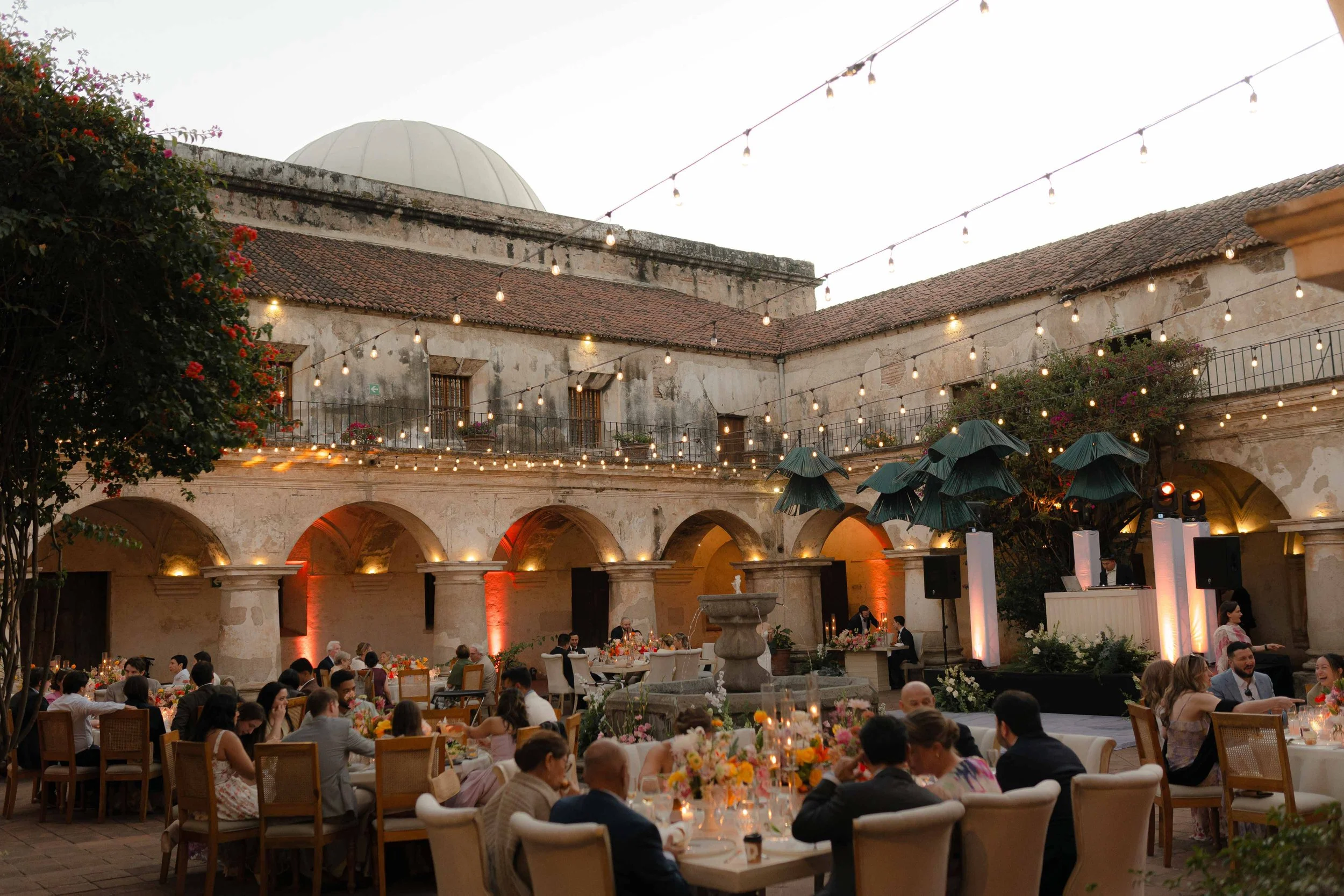 Una celebración en un patio con mesas decoradas con flores y velas, luces colgantes y un escenario con DJ, en el fondo una estructura antigua y un domo.