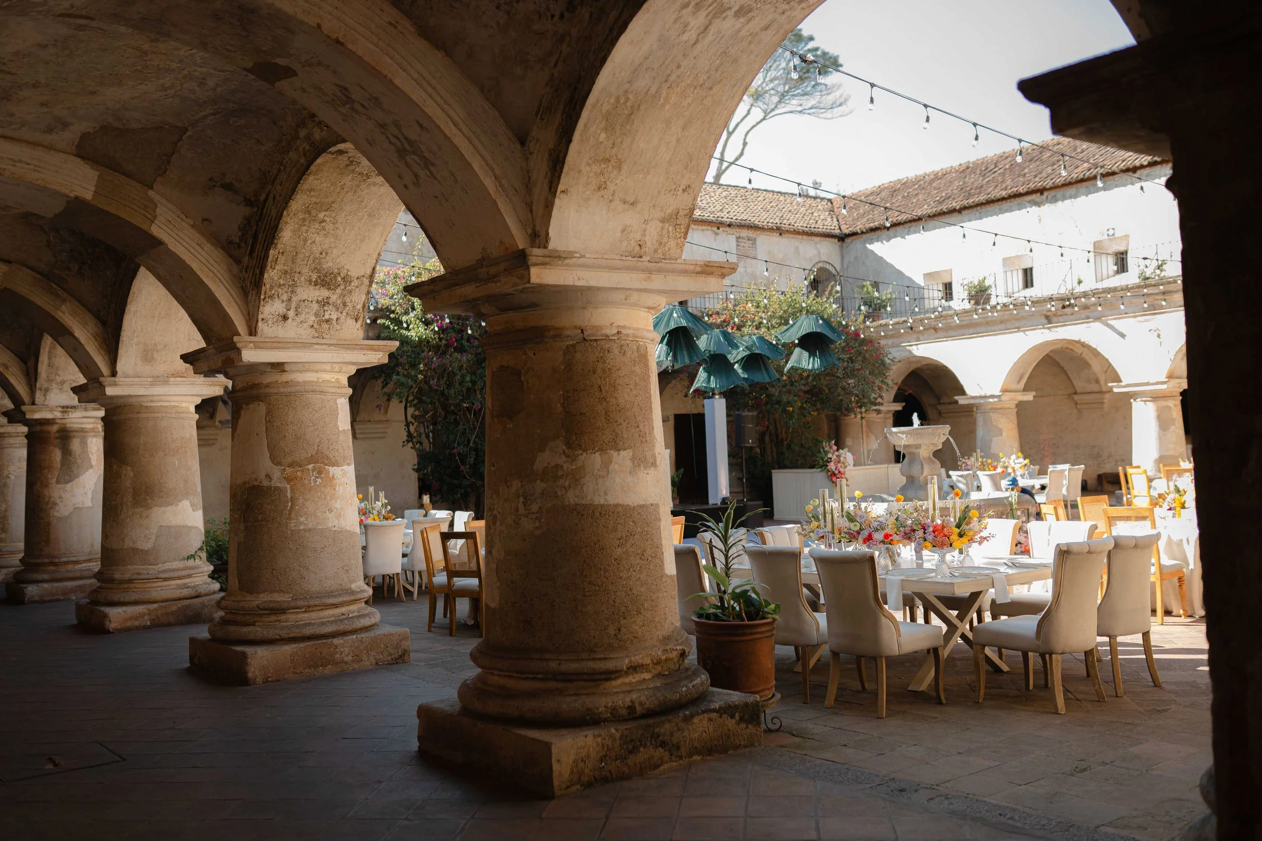 Patio interior con columnas de piedra, mesas decoradas con flores y sillas, decoraciones verdes colgantes y luces, en un ambiente al aire libre con edificio de techo de teja en el fondo.