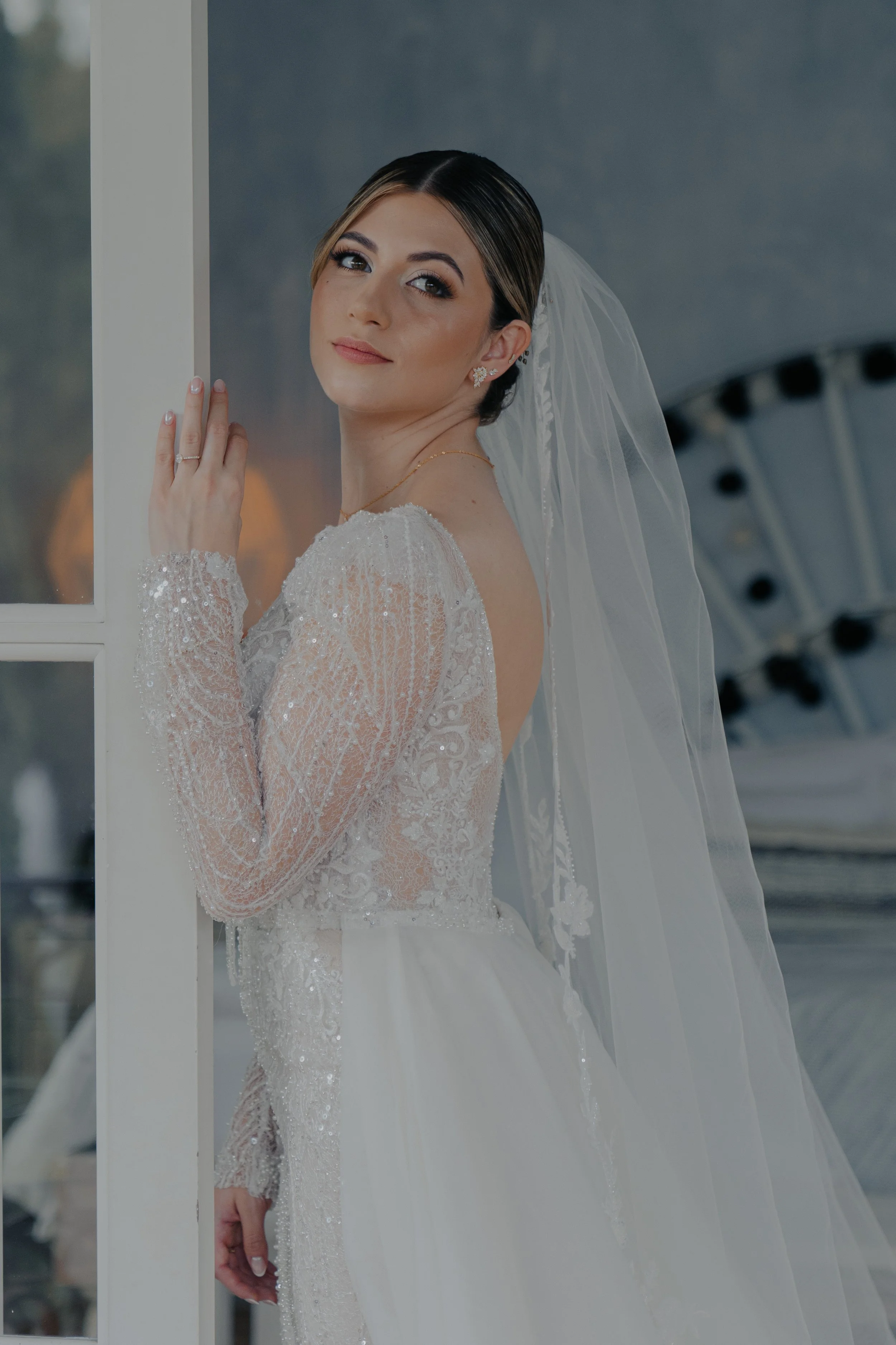 Mujer vestida de novia mirando hacia la cámara, con vestido de encaje y velo, posando junto a una ventana.