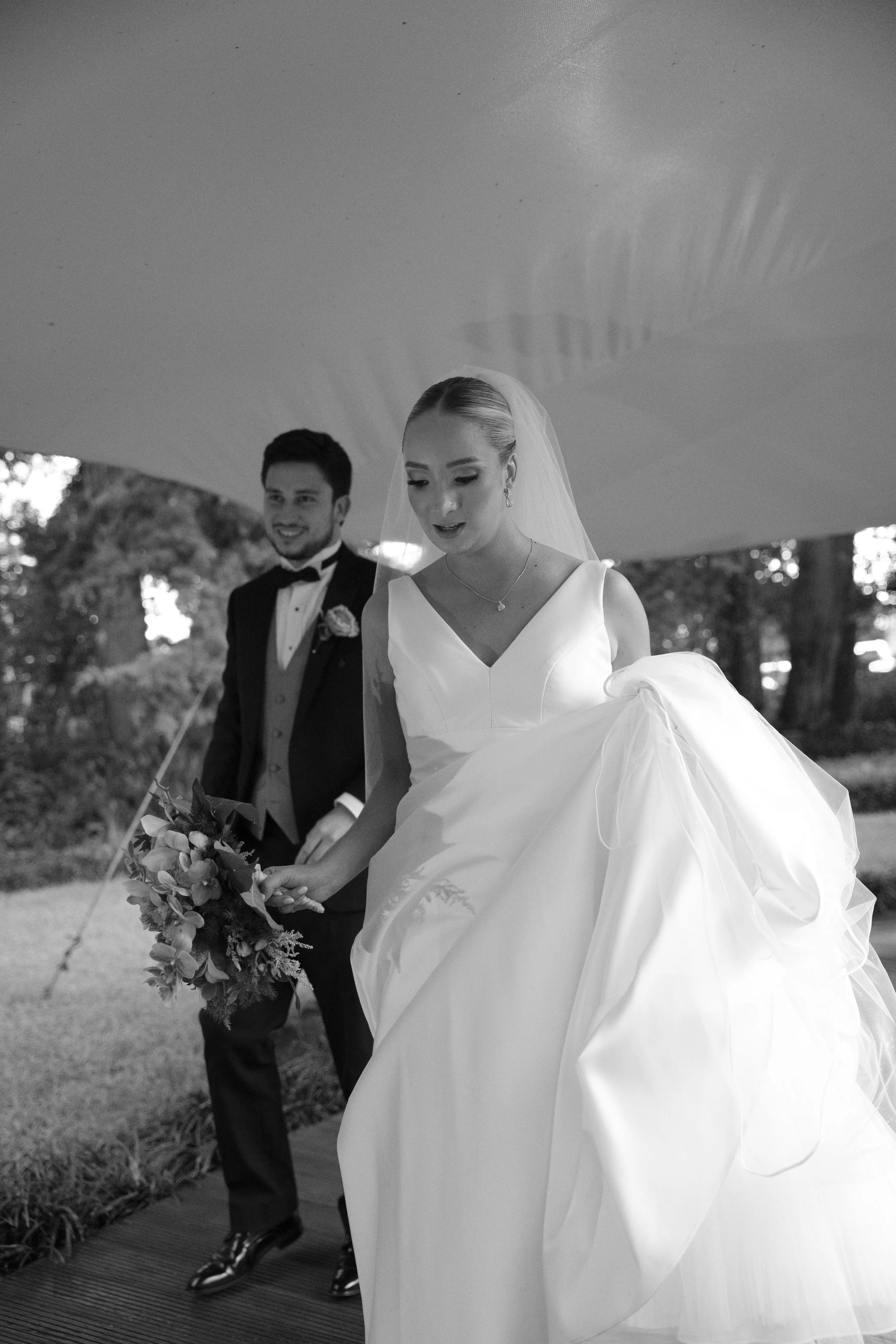 Una pareja de novios en su boda, la novia con vestido blanco y velo, la novia con ramo de flores y el novio con esmoquin, caminando por un entorno al aire libre.