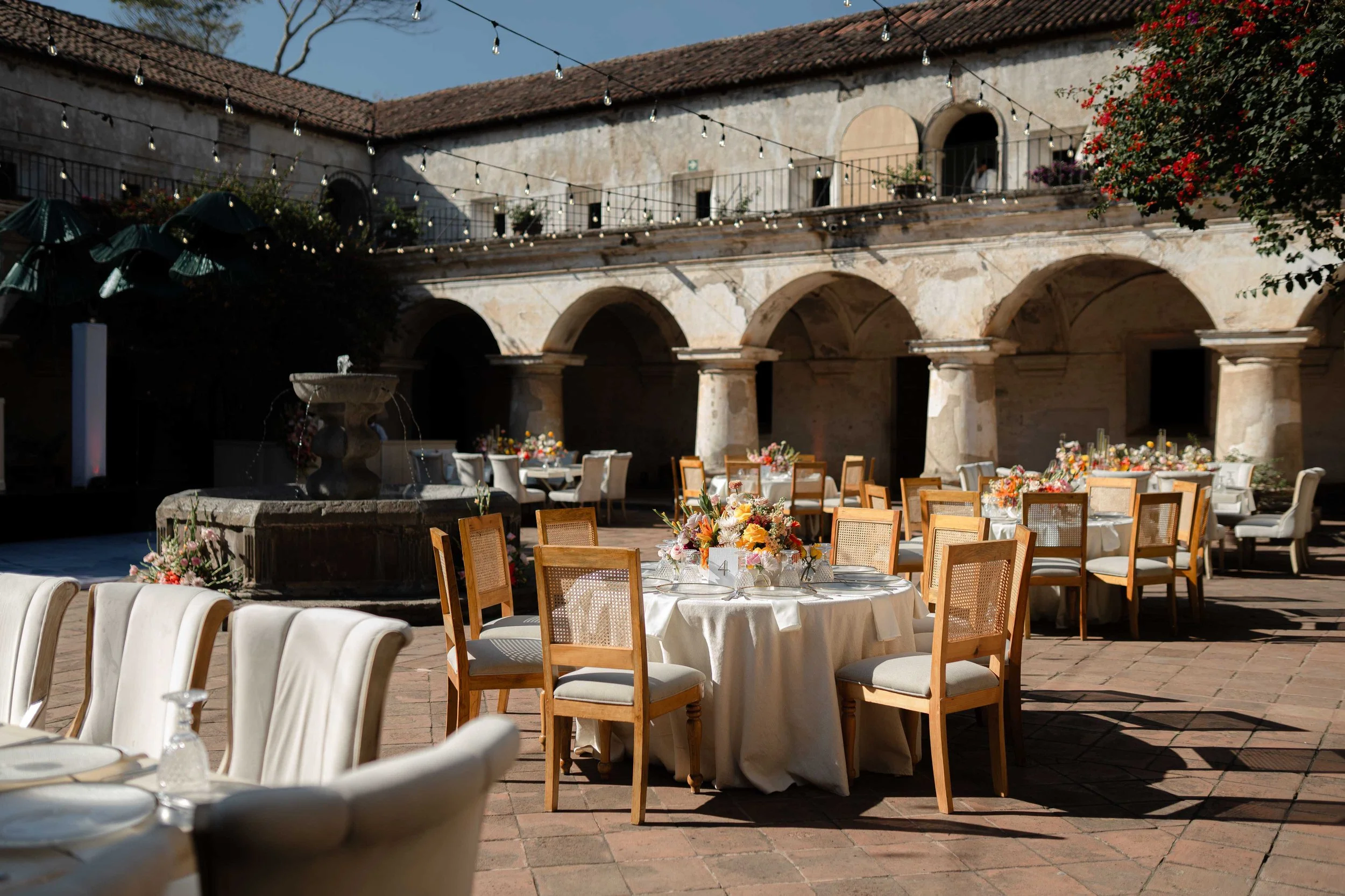 Una fiesta al aire libre con mesas decoradas con manteles blancos, centros de flores y utensilios de comida, en un patio con paredes de piedra, arcos y una fuente en el centro, decorada con luces colgantes.