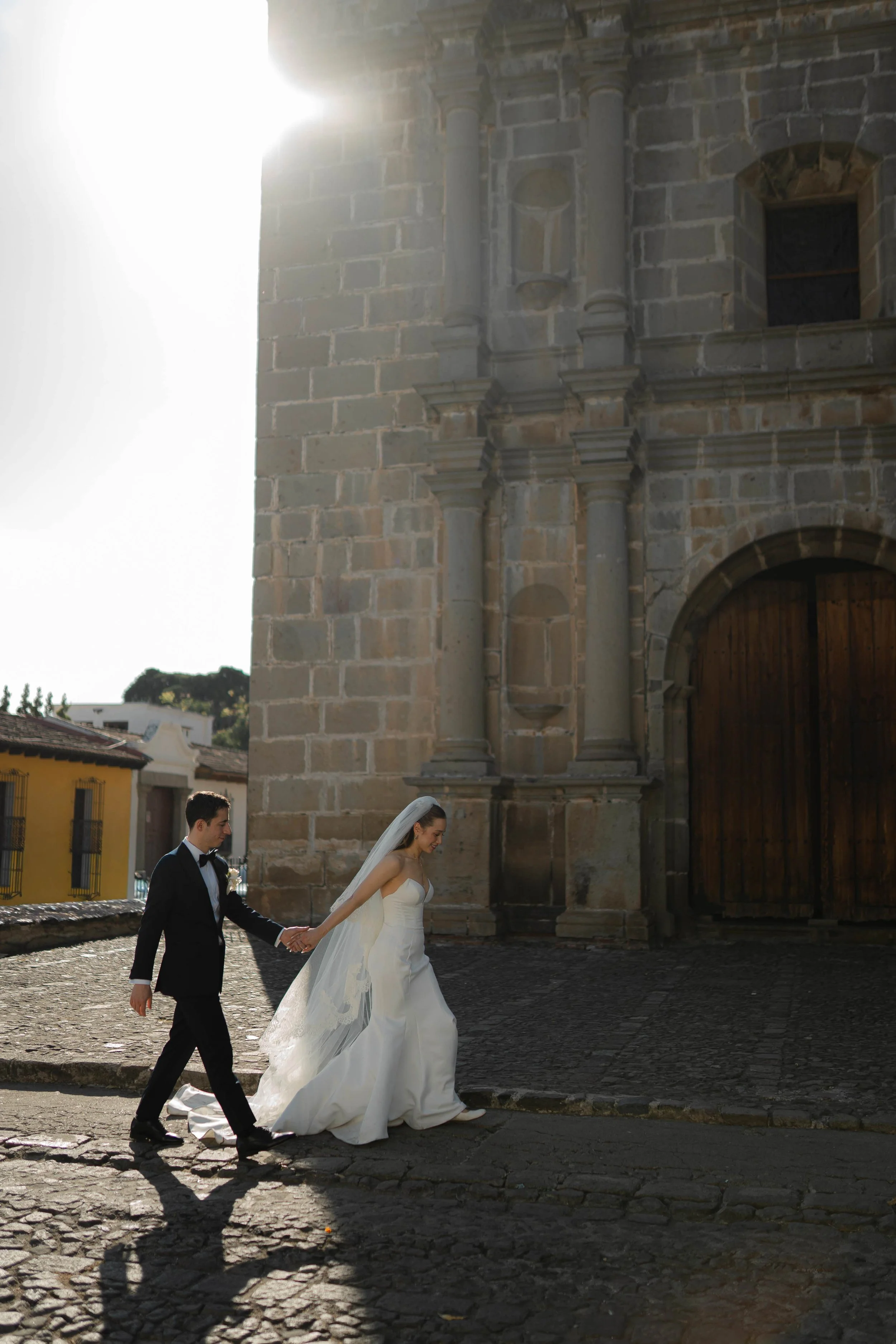Una pareja de novios camino frente a una iglesia antigua, con la novia usando un vestido de novia y el novio con smoking, en un día soleado.