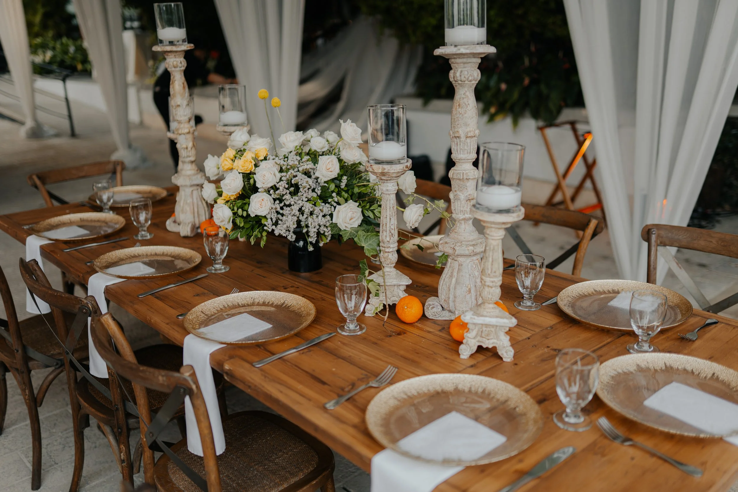 Mesa decorada para evento con flores blancas y amarillas, candeleros viejos y velas en frascos de vidrio, platos dorados, vasos de agua y cubiertos blancos y plateados en un ambiente elegante y acogedor.