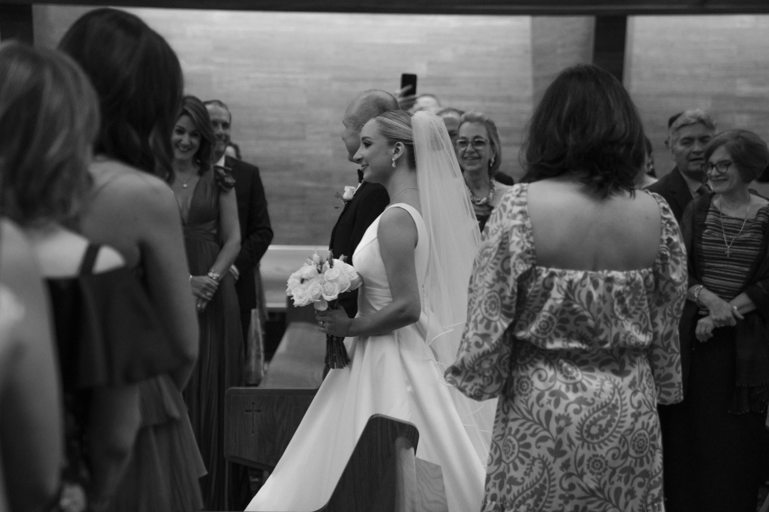 Una novia con vestido blanco y corona de flores en una ceremonia de boda, rodeada de invitados en un espacio interior
