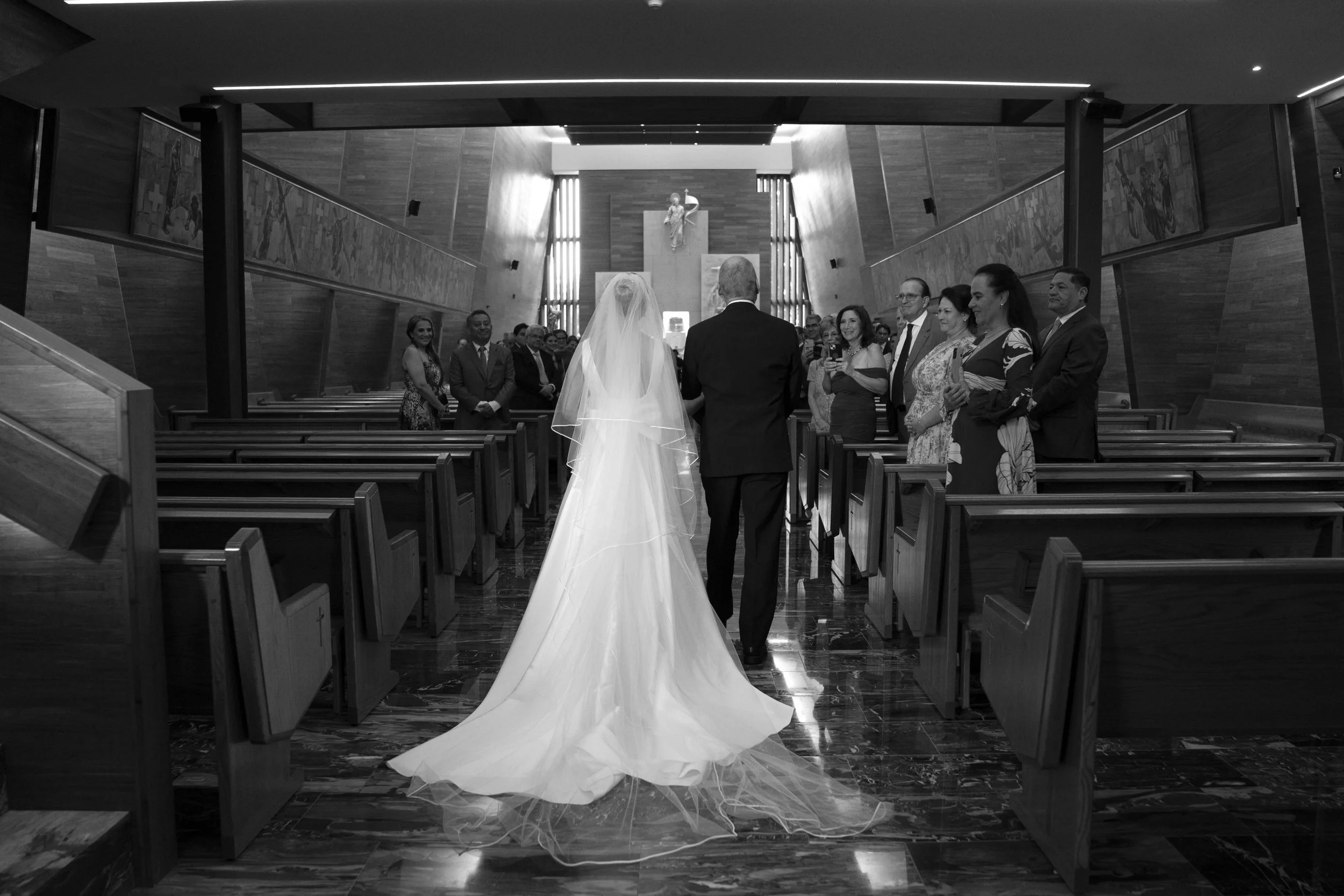 Una mujer vestida de novia caminando por el pasillo central de una capilla, acompañada por un hombre, con invitados observando y tomando fotos.