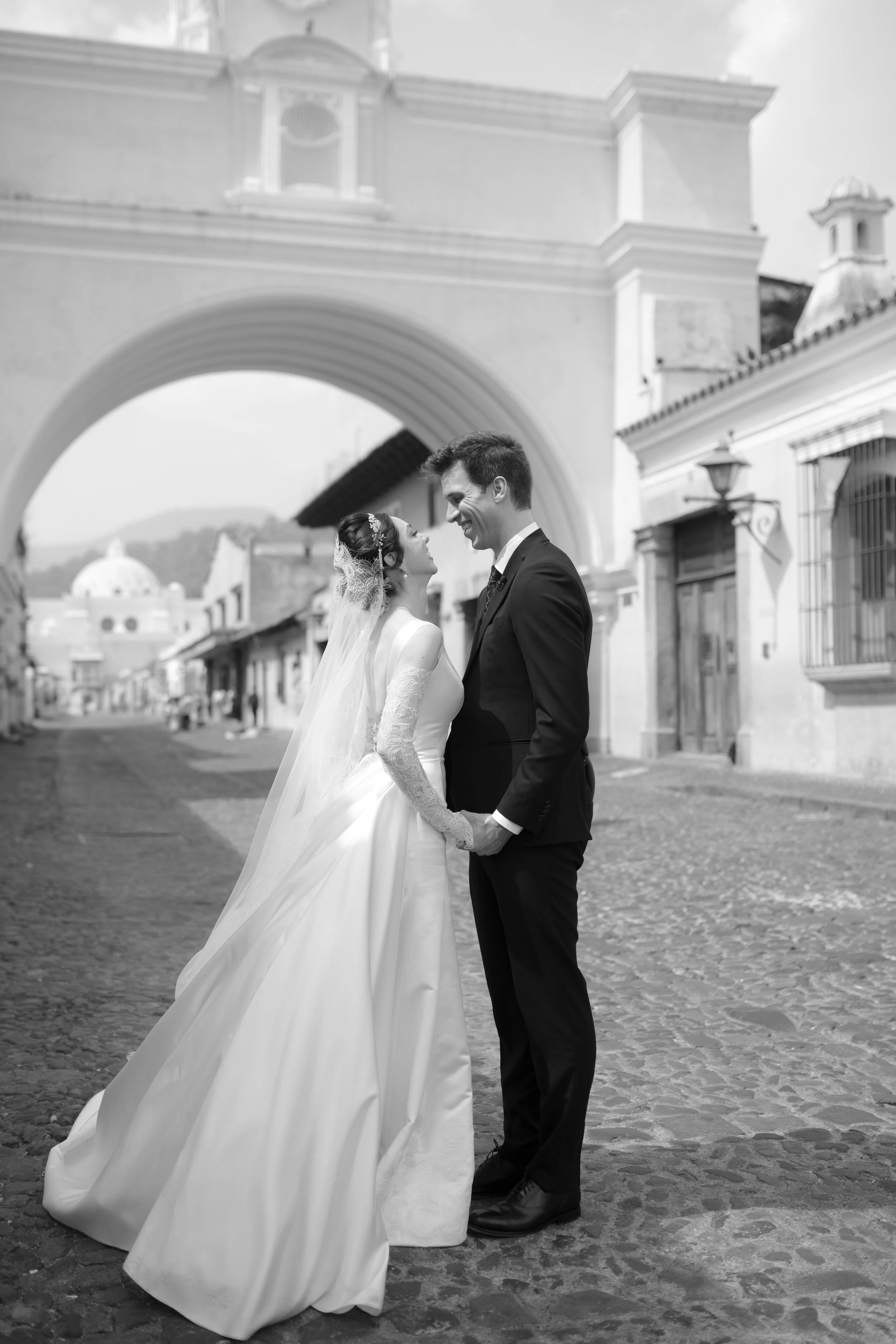 Pareja de recién casados en una calle empedrada, abrazados y sonriendo, con un arco y edificaciones antiguas en el fondo, en una fotografía en blanco y negro.