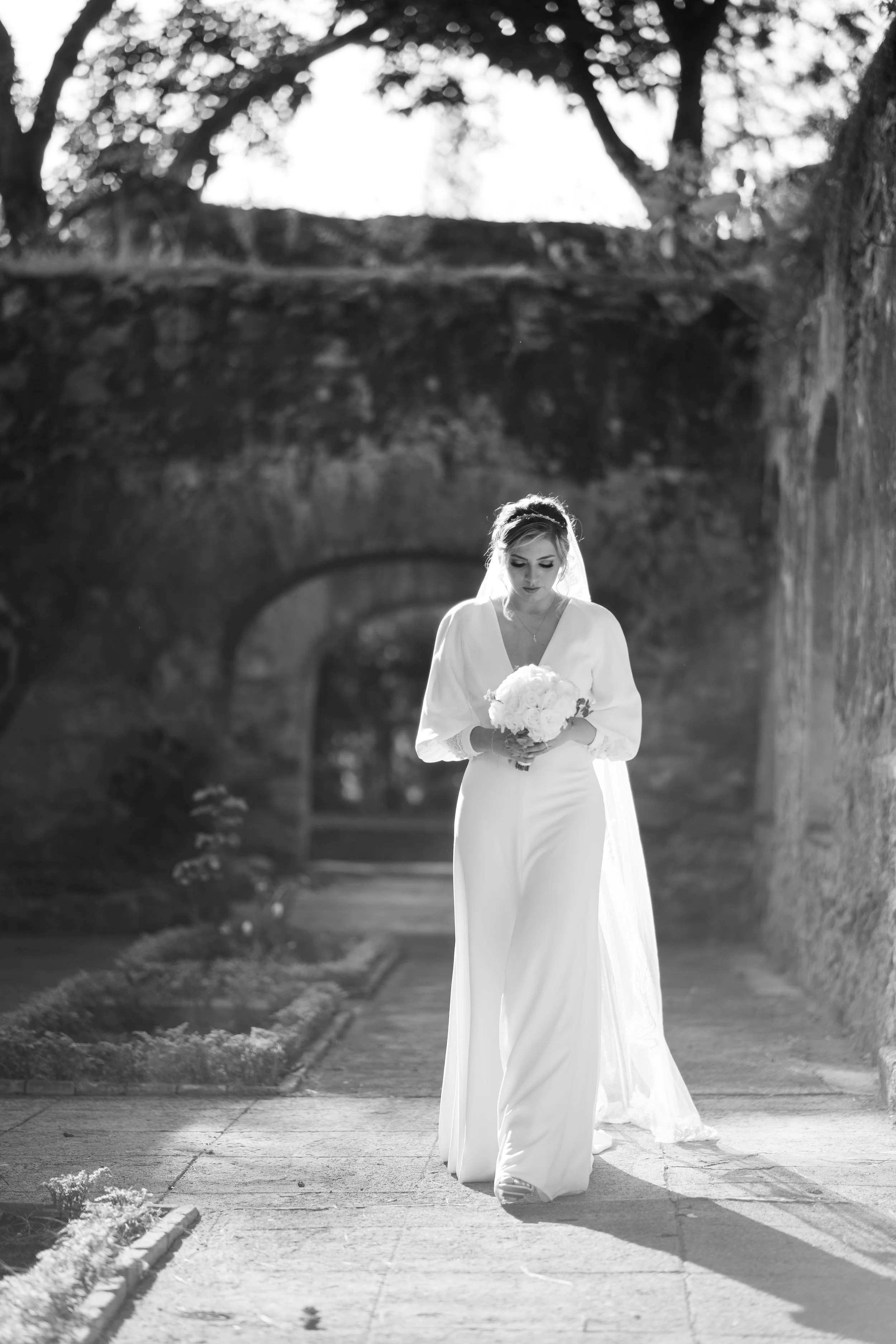Mujer vestida de novia caminando por un camino en un entorno natural, sosteniendo un ramo de flores, en una fotografía en blanco y negro.